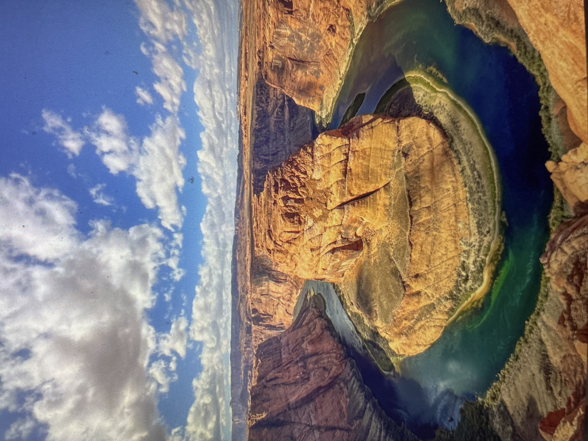 The image displays a high-angle, panoramic view of a dramatic natural landscape, prominently featuring a large river carving a distinctive, nearly circular bend through a deep canyon. This iconic geological formation is known as Horseshoe Bend, characteristic of the Colorado River.
The river itself exhibits varying shades of blue and green, indicating depth and water clarity, and is bordered by narrow strips of lighter, sometimes greenish, land along its banks. The surrounding canyon walls are composed of massive, stratified sandstone, displaying rich hues of orange, red, and brown, illuminated by direct sunlight. These rugged cliffs rise steeply from the river, showcasing intricate patterns of erosion and geological layers. A large, relatively flat-topped landmass forms the central 'island' around which the river loops.
Above the canyon, the sky is a bright, clear blue, adorned with scattered white cumulus clouds of various sizes. The weather appears sunny and pleasant, with clear visibility. The lighting suggests it is daytime, possibly late morning or early afternoon, with shadows adding depth to the canyon's features. There are no people, animals, or man-made structures visible within the main scene, emphasizing the untouched natural beauty and grandeur of the landscape. No text is visible in the image.