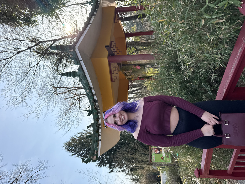 The image captures a young adult female standing on a red railing within the Zagreb Zoo in the City of Zagreb, Croatia. She has wavy, shoulder-length hair dyed in purple and pink ombre, wears a burgundy off-the-shoulder ribbed crop top, black trousers, and holds a burgundy handbag with "LIU JO MILANO" branding. Sunglasses are perched on her head as she smiles, looking towards the left. In the background, an Asian-style gazebo featuring a yellow base, red structural elements, and a prominent green, curved, ornate roof is partially obscured by dense green bamboo foliage. To the left, a wooden sign displays the text "CRVENI PANDA" (Red Panda) alongside images of red pandas. The environment includes numerous bare deciduous trees and some evergreen trees under a clear blue sky, with bright sunlight causing a lens flare in the upper right quadrant.