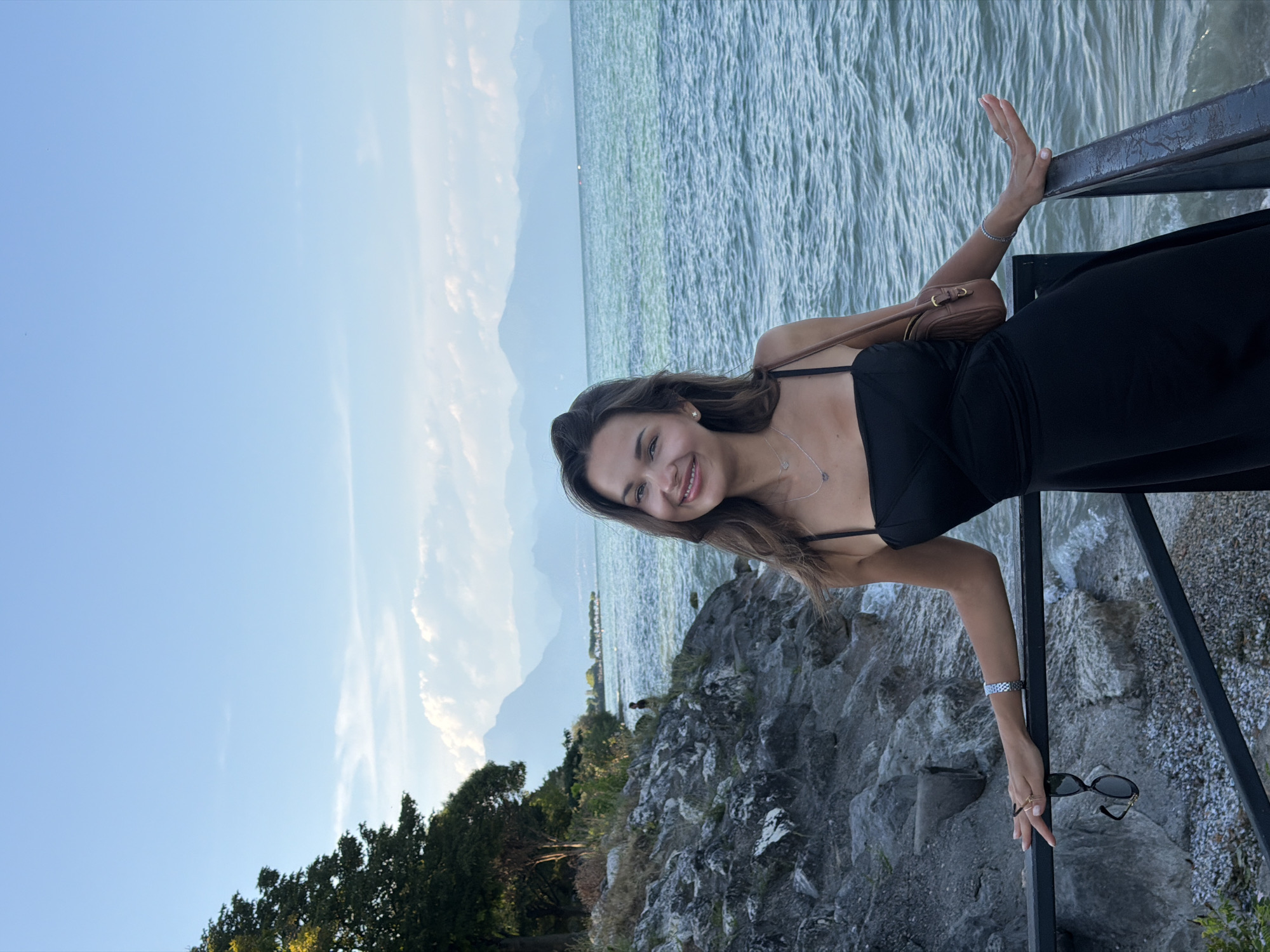 A medium shot captures a woman with long brown hair and a bright smile standing outdoors by a body of water. She is wearing a black dress with thin straps and a small brown purse slung over her right shoulder. She has a silver bracelet on her left wrist and is wearing two rings on her left hand. Her left hand rests on a black railing, while her right hand is extended slightly outwards. A pair of sunglasses hangs from the railing. The background features a rocky shoreline with some trees, a calm blue body of water, and distant mountains under a clear blue sky with scattered white clouds. The woman appears to be in Sirmione, Italy, likely near Lake Garda, given the landscape.