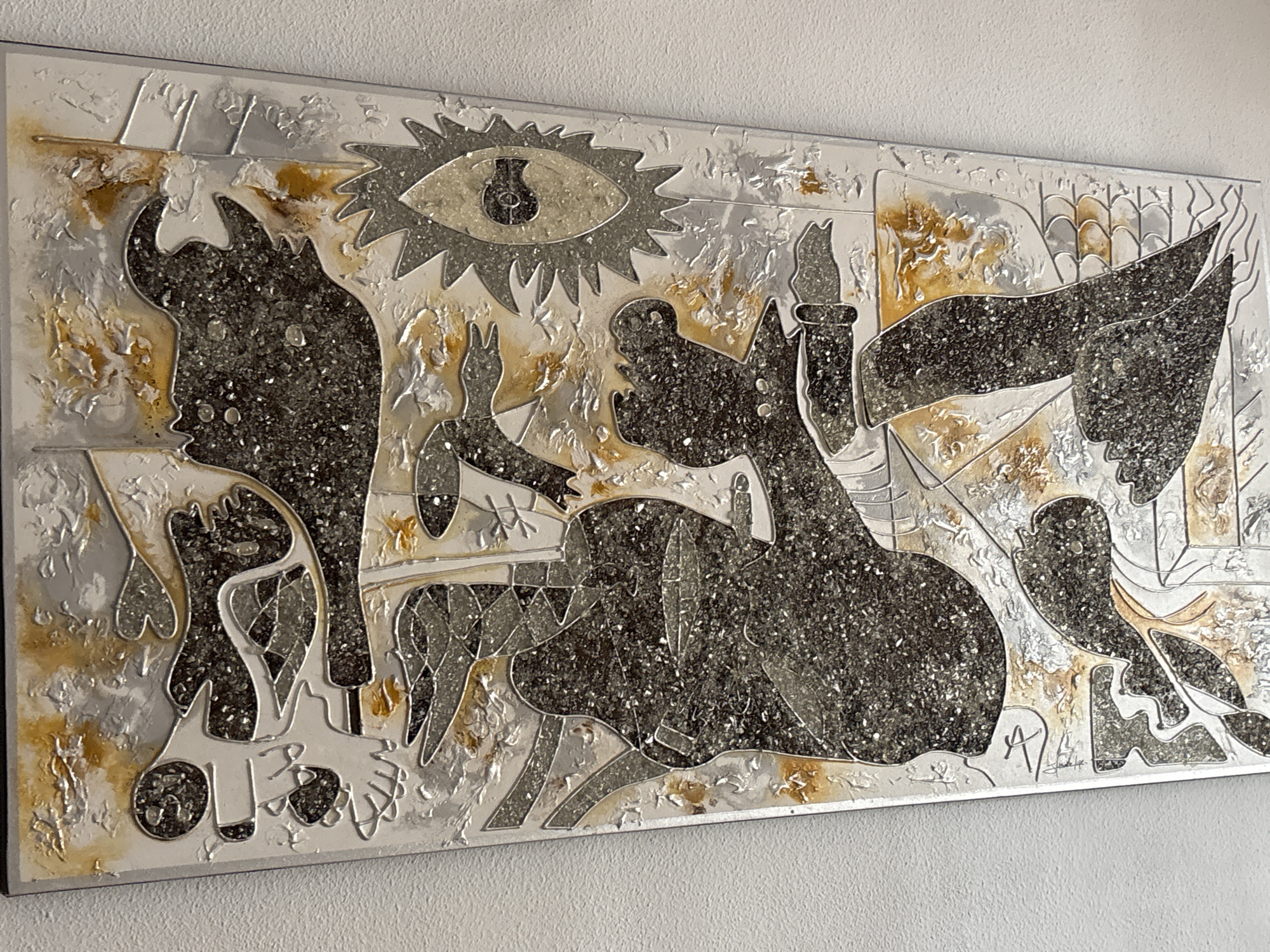 This is a textured, abstract artwork featuring stylized figures and symbols in shades of silver, black, and gold. The composition is rectangular and appears to be mounted on a white wall. On the left, a dark, jagged silhouette resembling a bull's head is prominent. In the center, an eye with a keyhole motif is positioned above several fragmented human-like forms. To the right, more abstract shapes suggest wings or fabric. The artwork has a metallic sheen and appears to incorporate glitter or small reflective particles within its darker sections, creating a textured surface. The overall style is reminiscent of cubism or surrealism, with sharp lines and distorted perspectives. A signature is visible in the lower right corner. The artwork is displayed indoors, on a textured white wall, which is characteristic of an art gallery or exhibition space. No specific location in Orihuela, Spain, can be determined from the visual information provided.