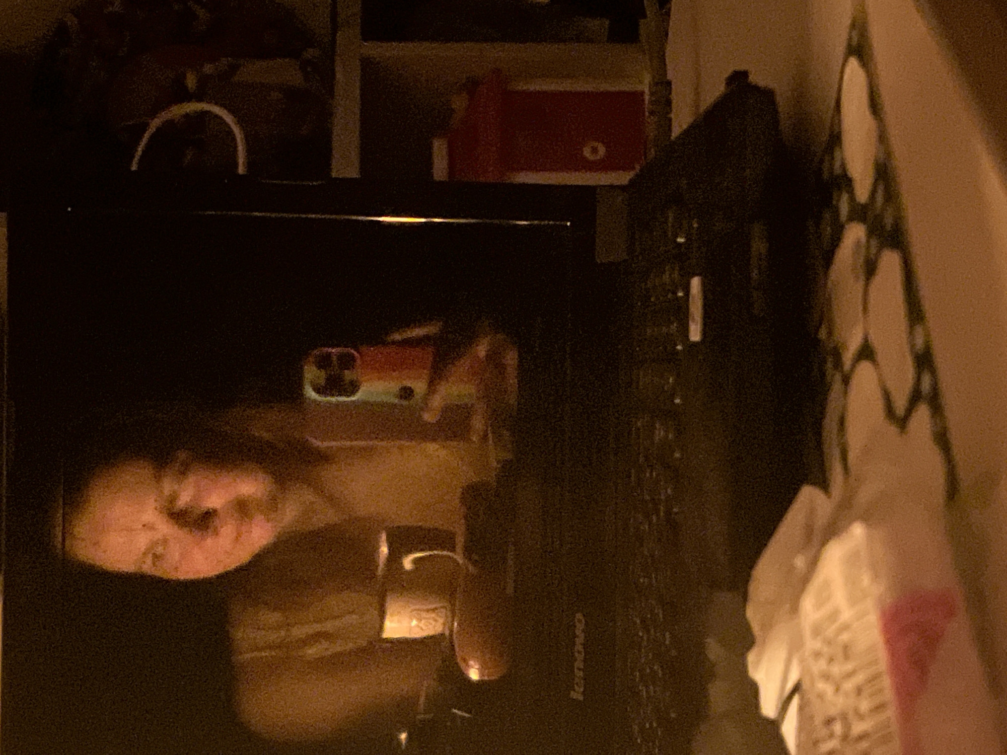 A blurry image shows a reflection of a woman holding a phone in front of a Lenovo laptop. The environment is dark and difficult to discern. There are shelves visible in the background, with various items including a red box. A box with white and pink designs is on the right side of the image. The location is unknown.