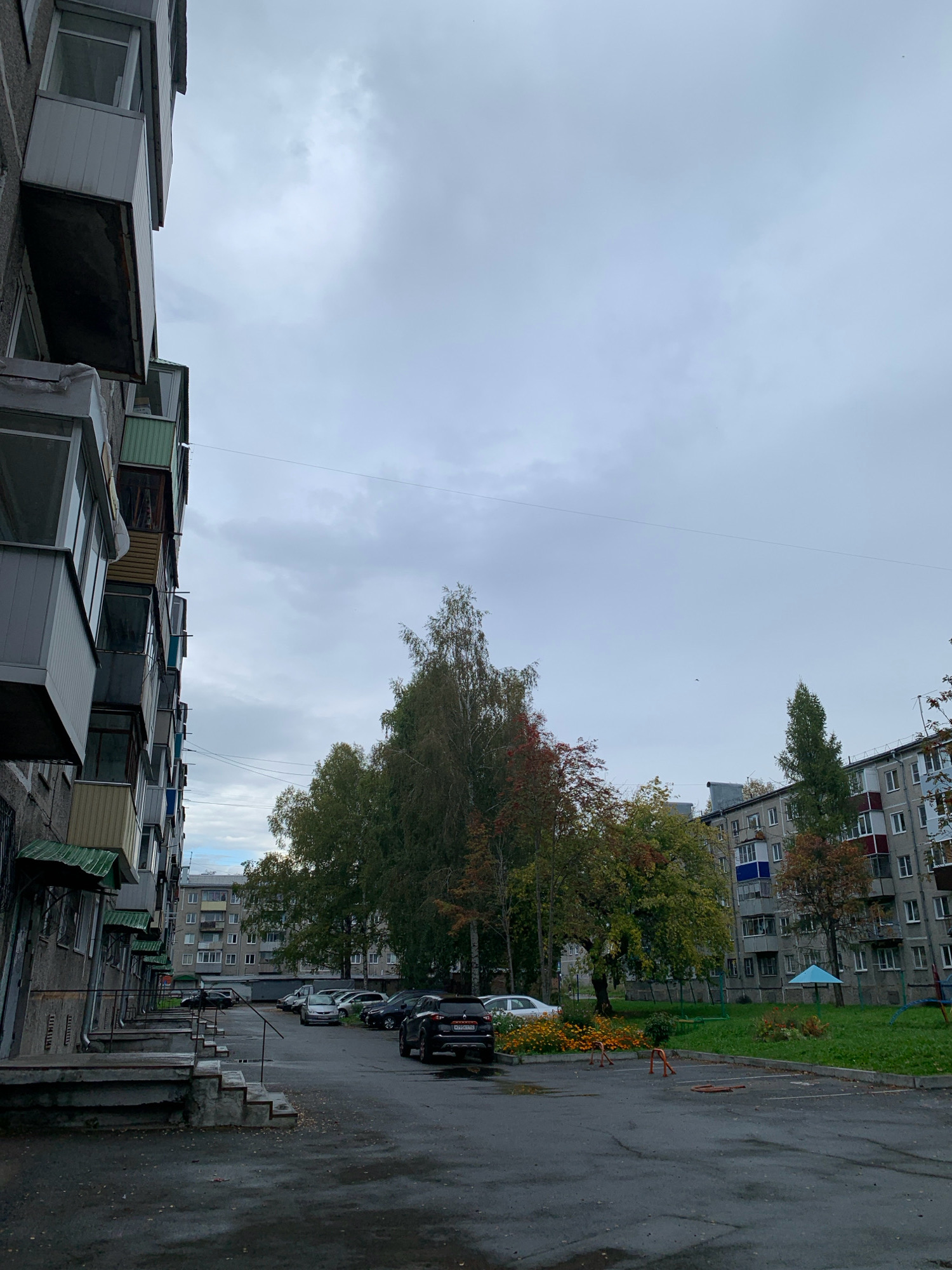 A vertical, medium shot captures a cloudy, overcast day in a residential courtyard in Prokopyevsk, Russia. On the left, the side of a weathered, multi-story apartment building dominates the frame, its balconies protruding uniformly. The building appears to be constructed from concrete. The ground is a wet asphalted courtyard with several parked cars, including a dark SUV in the foreground. Mature trees with a mix of green and autumn-colored foliage are scattered throughout the courtyard, with a cluster of trees forming a significant visual element in the mid-ground. In the background, another apartment building, also multi-story, is visible. A small, light-blue playground umbrella is set up on a grassy area to the right. The overall atmosphere is quiet and still, suggesting an ordinary day in an urban residential area.