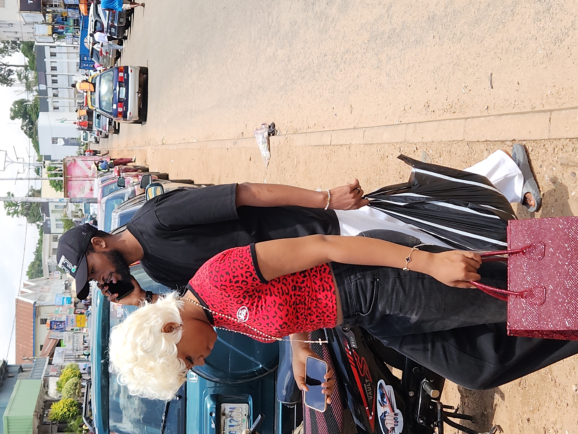 Two people stand near a road in what appears to be a city. The person on the left is a woman with blonde, curly hair wearing a red leopard-print crop top, black pants, and a rosary necklace with a cross. She is holding a smartphone in her left hand and a red, textured handbag with handles in her right. The person on the right is a man wearing a black t-shirt and a black baseball cap with a logo. He is holding a black smartphone to his right ear and speaking on it. He is also holding a black and white striped umbrella in his left hand. The background shows a street lined with cars and buildings. The road is paved, and there is a concrete curb. Some debris is visible on the side of the road.