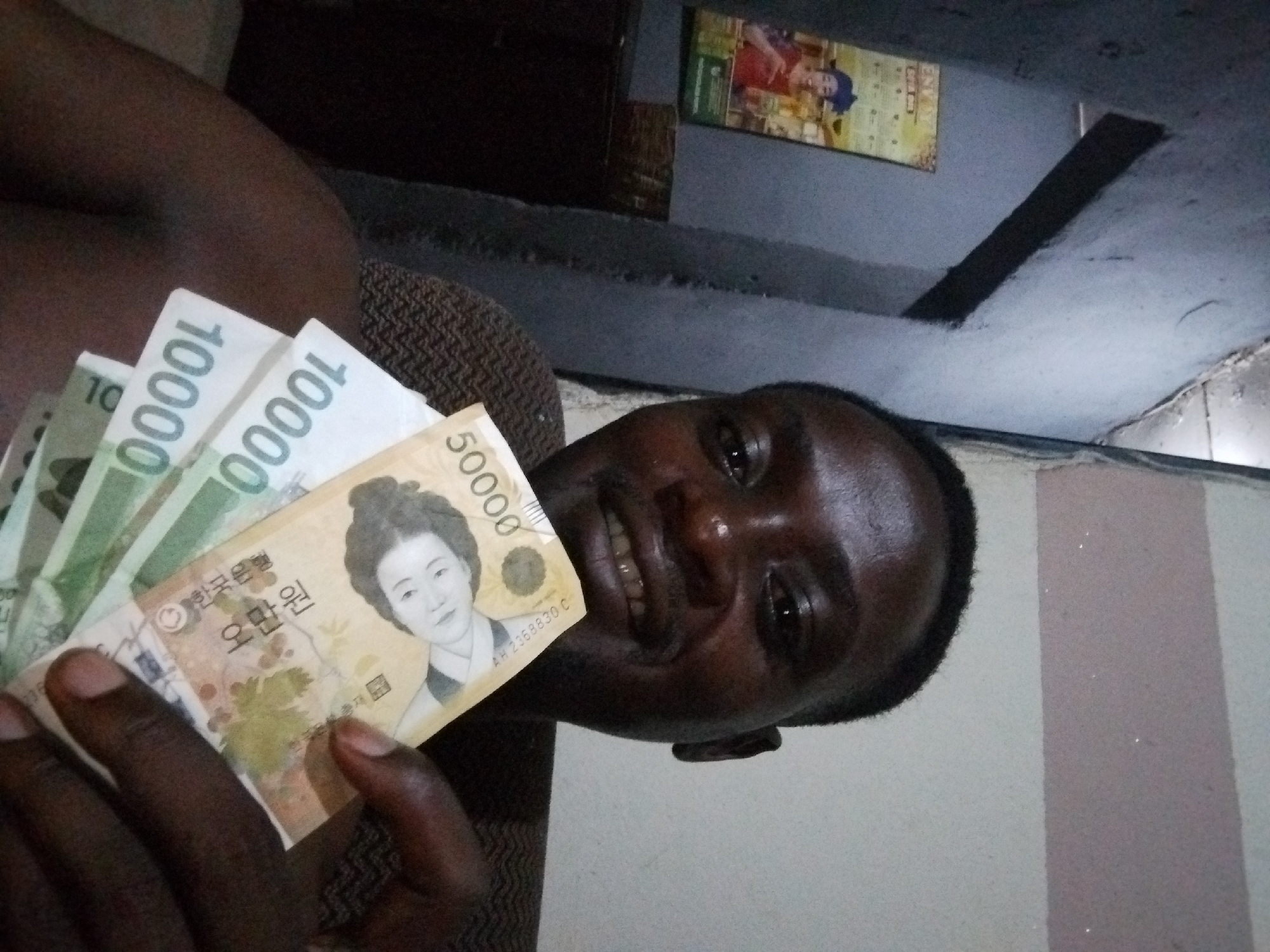 A man is displaying Korean currency bills (10,000 and 50,000 Won denominations) in an indoor environment. The location appears to be a room with a poster on the wall and a visible ceiling structure. The precise city and country are undetermined.
