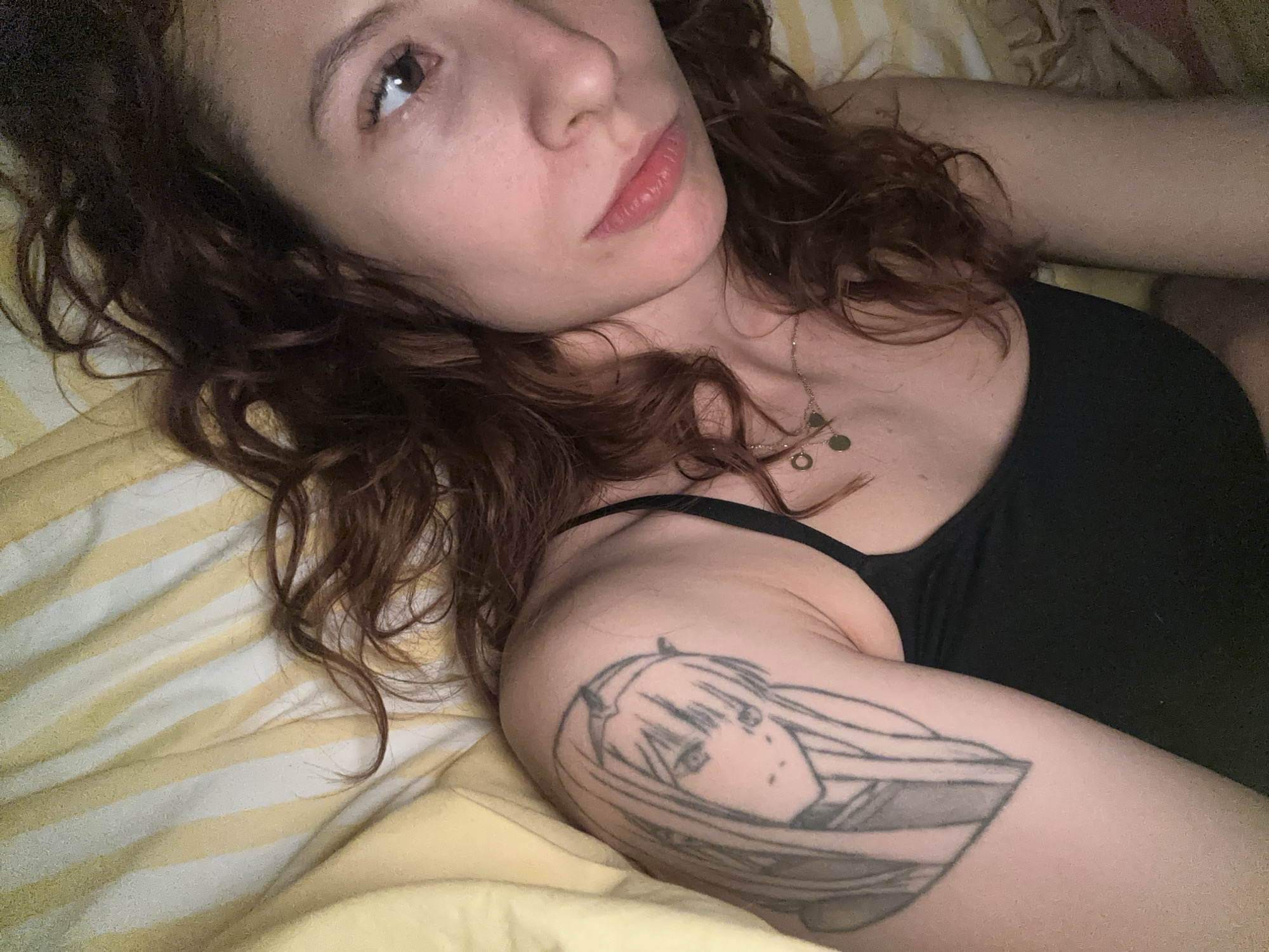 A woman with red curly hair lies on a bed with a yellow and white striped blanket. She is wearing a black tank top and a necklace with three coin-like pendants. There is a tattoo of a manga character on her left upper arm. She is looking upward and to the left. The location is not discernible from the image.