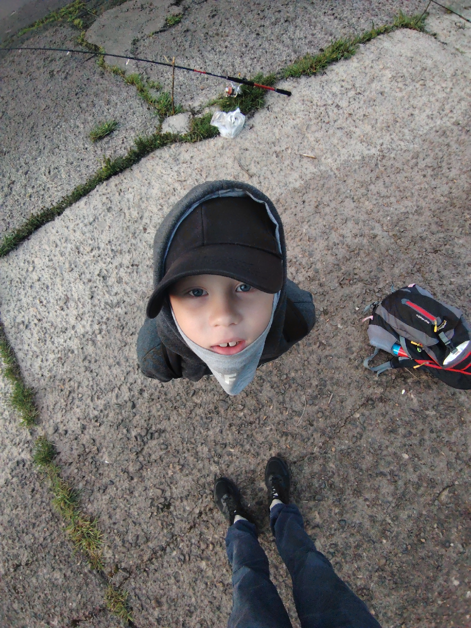 A high-angle shot captures a young child looking up at the camera, with an adult's legs and feet visible at the bottom of the frame. The child, with fair skin and blue eyes, wears a dark gray hooded sweatshirt, a black baseball cap, and a light gray neck gaiter pulled up to his nose, revealing a faint, unreadable letter or logo, possibly an "F". To the right of the child, a black and gray backpack with red and yellow accents lies on the ground; text on it is too blurry to discern.
The setting is outdoors in Chaikovsky, Russia, on a textured ground of light-colored concrete or asphalt. Cracks in the pavement are filled with green grass and weeds, indicating a somewhat worn or naturalized surface. In the upper left corner, a fishing rod with a reel is laid on the ground. The presence of the fishing rod and backpack suggests a recreational activity, likely fishing or an outing. The lighting is soft and diffused, possibly indicating an overcast day or the softer light of early morning or late afternoon. The child's expression is neutral and direct, engaging with the person taking the photo.