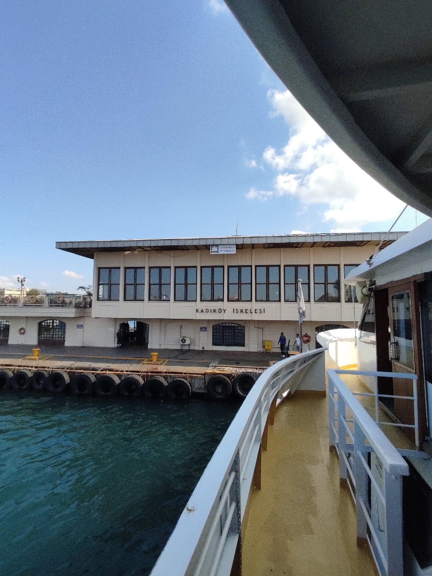 This image depicts the Kadıköy ferry terminal in Istanbul, Turkey, seen from the deck of a ferry. The terminal building is a multi-story structure with a white facade, featuring a prominent sign that reads "ISTANBUL" in a logo and "KADIKÖY İSKELESİ" below. Large, dark-framed windows line the upper level of the building. In front of the building, a dock area is visible, with a line of large, black tires acting as fenders along the edge. The water of the Marmara Sea is a deep teal color and appears slightly rippled. On the right side of the image, the railing and deck of the ferry are visible, painted white and yellow respectively. Two individuals, seemingly ferry workers or passengers, are present near the entrance of the terminal, one wearing a blue uniform and the other a white shirt. The sky is a clear blue with a few scattered white clouds.
