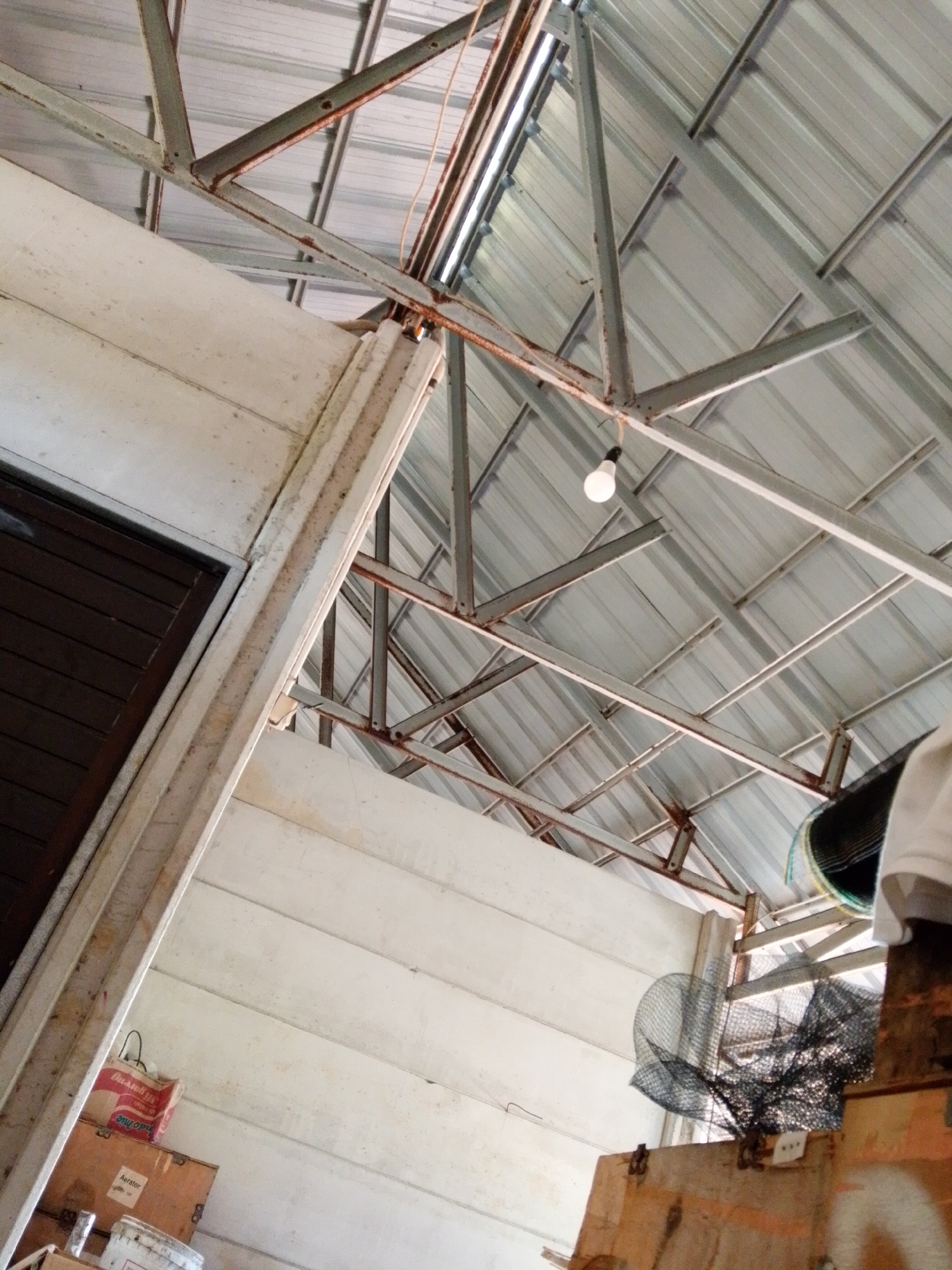 The image, captured in Bukit Ketok, Indonesia, showcases the interior of a structure with a corrugated metal roof supported by a steel frame. A single light bulb hangs from the frame. To the left, a wall constructed of horizontal planks is partially visible, with a dark-colored door. Several boxes and containers are stacked on the floor near the wall. To the right, a black netting-like object is positioned near a wooden structure.