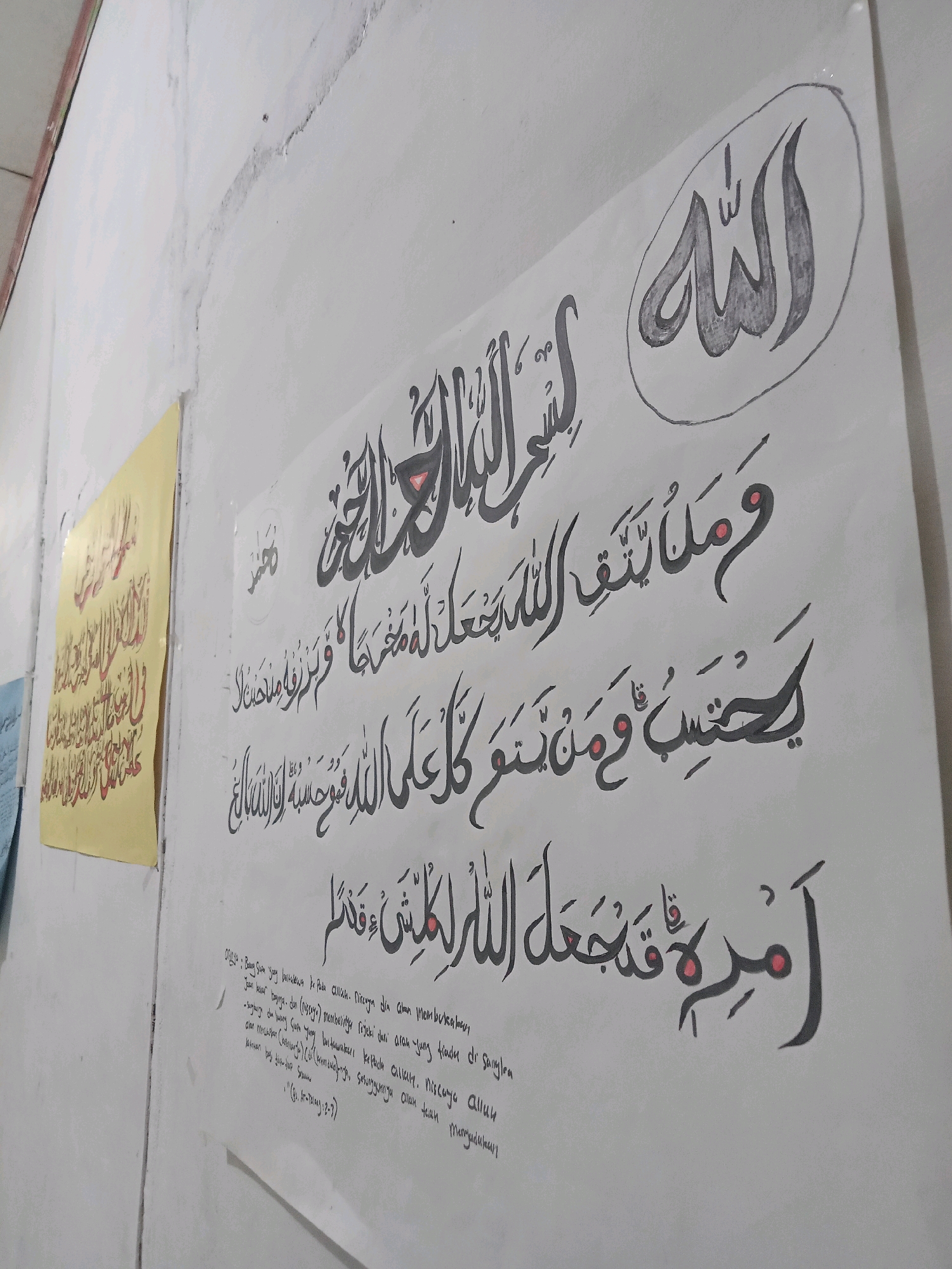 The image displays a close-up view of a white wall in what appears to be an interior space. On the wall, there is a large, unframed piece of paper featuring Arabic calligraphy. The calligraphy is executed in black ink with some red accents. At the top right, the word "Allah" is written in a circular design. Below it, there are several lines of Arabic text. To the left of this main calligraphy piece, there is a smaller, yellow paper with red calligraphy, also affixed to the wall. A portion of a light-colored ceiling or upper wall structure is visible at the top of the image. The overall impression is that of a decorated wall, possibly in a religious or educational setting. No people or specific location details beyond "Sukomananti, Indonesia" are discernible from the image itself.