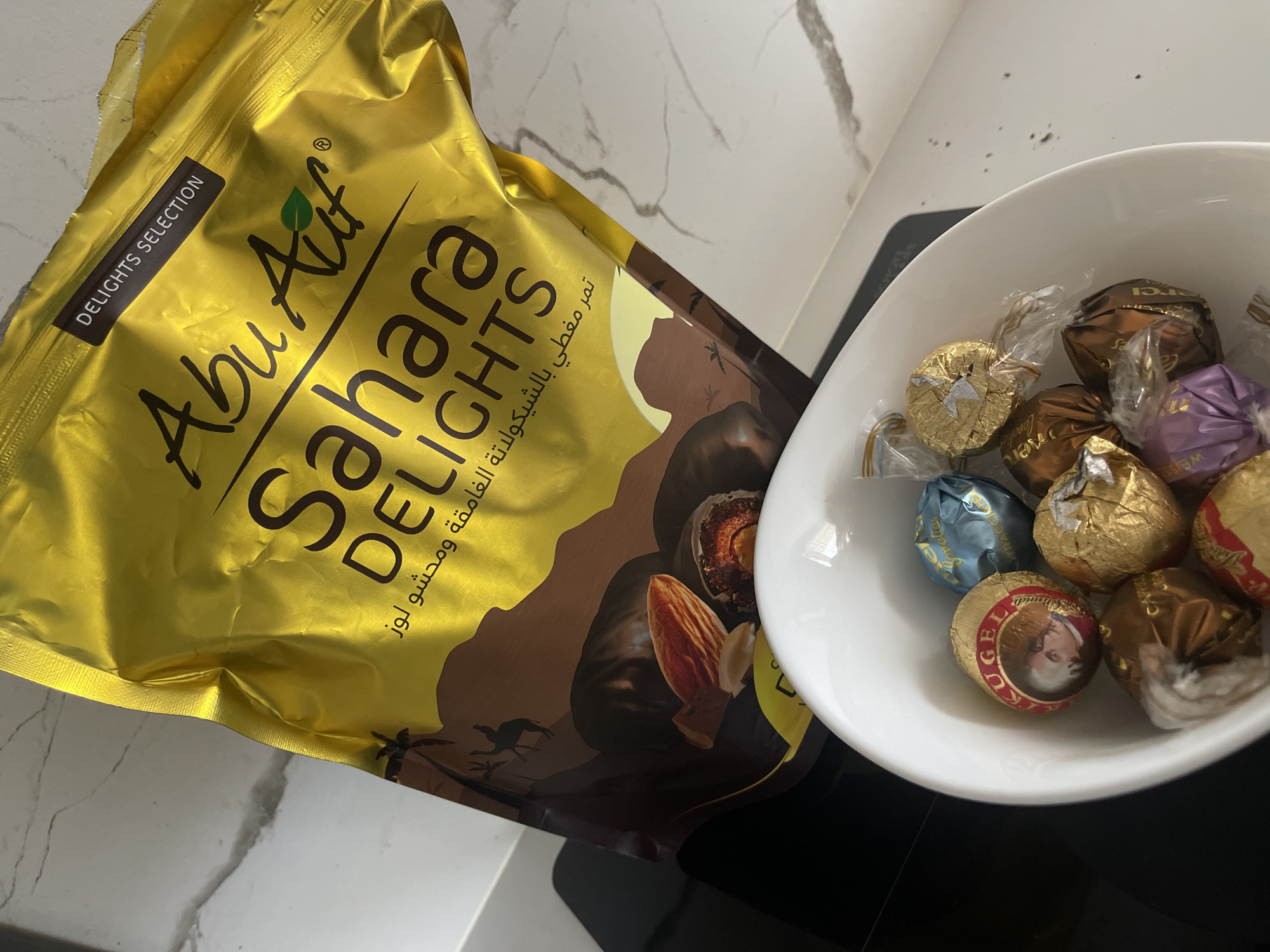 The image presents a bag of "Abu Auf Sahara DELIGHTS" next to a white bowl filled with individually wrapped chocolate candies. The bag is gold with brown accents, depicting dates and almonds, and includes text in Arabic. The candies in the bowl are wrapped in various metallic colors, including gold, brown, blue, and purple, some with discernible labels. The scene is set on a white and gray marble surface, suggesting a kitchen or dining area. There is no indication of Sofia, Bulgaria, or any people within the image.