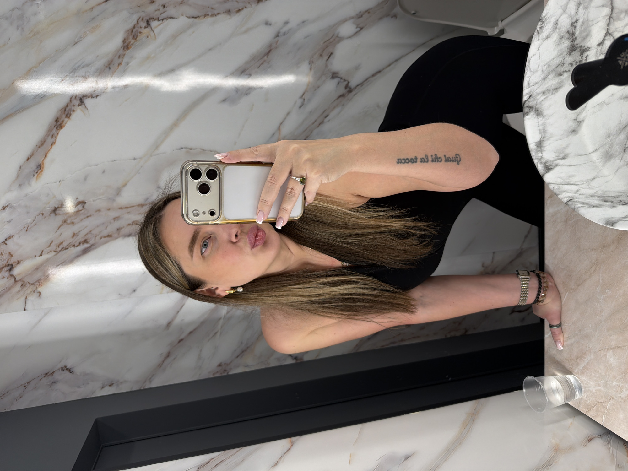 A woman is captured taking a mirror selfie in what appears to be a modern and elegantly designed bathroom or restroom, located in Moscow, Russia. She holds a gold-colored smartphone in her right hand, positioned to reflect herself. Her lips are slightly pursed, and she looks directly into the phone's camera with a focused expression.
She has long, straight hair with blonde and brown highlights, falling over her shoulders. She wears a black top or bodysuit. On her right forearm, a script tattoo is visible, which reads "Gual la Rocca" when viewed in the mirror, indicating the text is mirrored. Her nails are neatly manicured with white French tips. A ring featuring a prominent green stone adorns her right ring finger. On her left wrist, she wears a silver-toned watch alongside several beaded bracelets. A pearl earring is visible in her left ear.
The setting is characterized by polished surfaces, likely marble or a marble-patterned material, featuring white and brown veining on the walls and countertops. A bright, horizontal light streak reflects across the upper wall. A dark gray or black counter or frame runs across the lower portion of the image. To the far right, a white and gray marble-patterned circular tray or dish holds a dark, undefined item. A clear disposable plastic cup containing water is placed on a lower counter surface. The environment suggests a clean and contemporary aesthetic.
