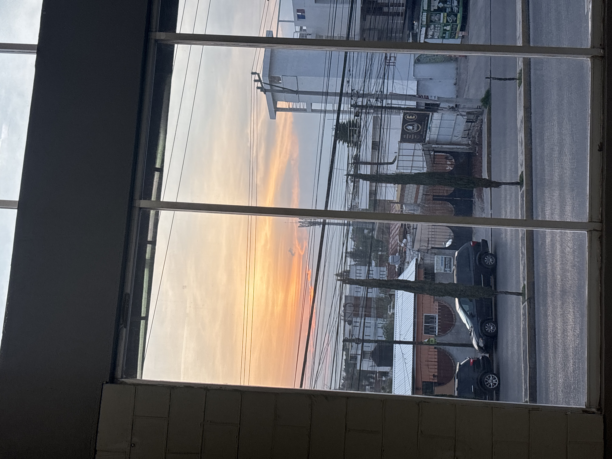 A view from a building in Puebla, Mexico, through a multi-pane window. The time of day appears to be either sunset or sunrise, judging by the colors in the sky. Outside the window are buildings, power lines, and a street. There are at least three cars parked on the street. One of the cars is black.