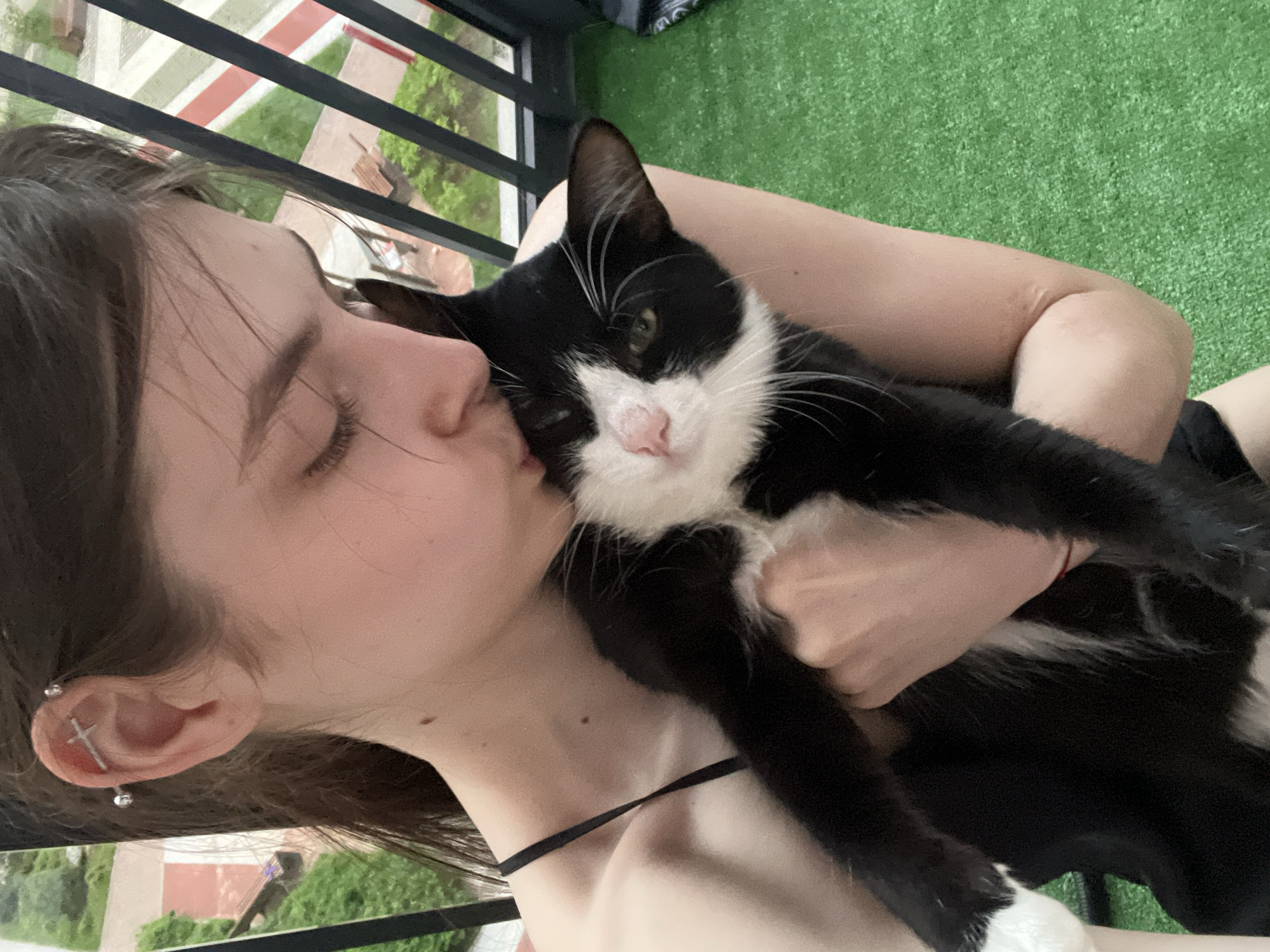 A young woman is holding and kissing a black and white cat outdoors. The woman has brown hair and is wearing a black choker necklace. She has a piercing in her ear shaped like a cross. The cat has white fur on its chest and paws. The background shows a balcony with green artificial turf. A railing in the foreground and other buildings in the background suggest an urban setting. It is not possible to determine if the location is Saint Petersburg, Russia without additional information.