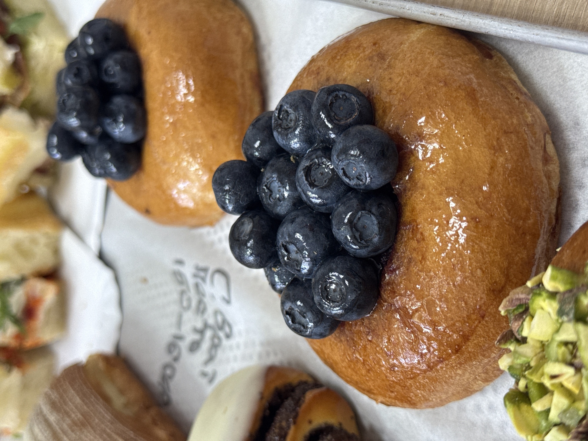The image captures a close-up, high-angle view of a selection of baked goods and pastries, likely displayed on a white paper liner within a light-colored tray. The main subjects are two golden-brown, glossy pastries, possibly brioche buns or donuts, each topped with a cluster of fresh, plump blueberries. The pastry on the right is in sharp focus, revealing the shine of its glaze and the intricate details of the blueberries.
Around these central items, other food elements are partially visible. To the far left, out-of-focus savory items, resembling small sandwiches or canapés with green garnishes, can be seen. In the bottom foreground, other pastries are partially shown: one with a white-frosted edge and a dark spiral filling, and another with a ribbed, light brown texture. To the bottom right, another pastry is visible, generously topped with finely chopped green nuts, likely pistachios.
The white paper lining beneath the pastries features blurred black text. While most of it is illegible due to the angle and blur, the numbers "1860-1960" are discernible. Other stylized letters are also visible, potentially Cyrillic, given the location context of Belgrade, Serbia. The lighting is bright and even, highlighting the fresh appearance and appealing textures of the baked goods. No people or specific events are depicted; the scene is a still life of a dessert and savory food display.