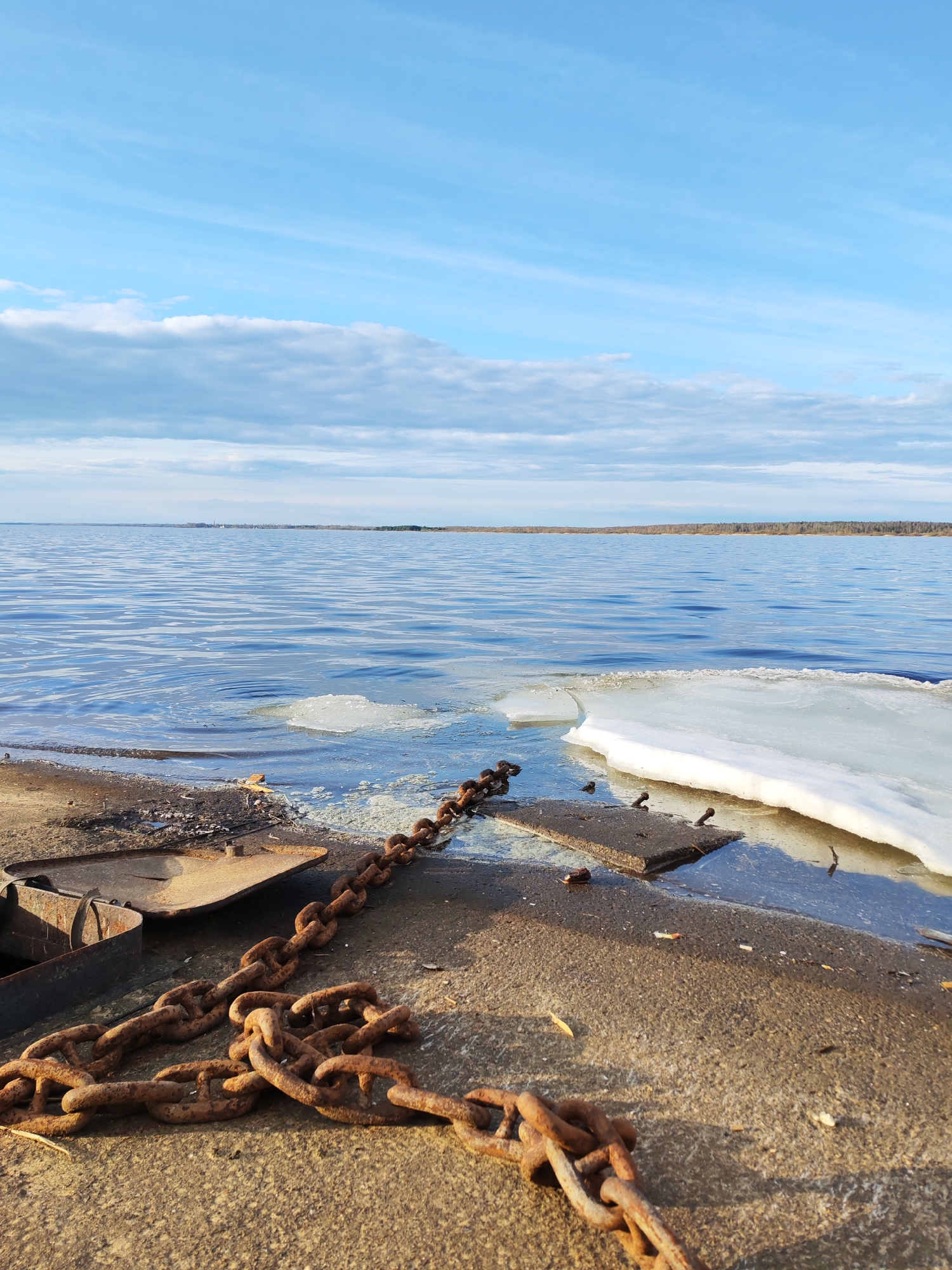 The image shows a waterfront scene, possibly a pier or embankment, in Rybinsk, Russia. The foreground is dominated by a heavy, rusted chain lying on a concrete surface, which appears to be part of the pier. Scattered pieces of what might be metal debris, including a partially visible metal box, are also present. In the immediate background, there is a partially frozen body of water, with ice patches floating near the shore. The water has a blue-grey hue. Beyond the water is a distant shoreline with a visible treeline. Above, the sky is partly cloudy with a mix of blue and white clouds. The lighting suggests it's daytime, likely in the late afternoon or early morning due to the low angle of the sun. There are no people visible in the image.