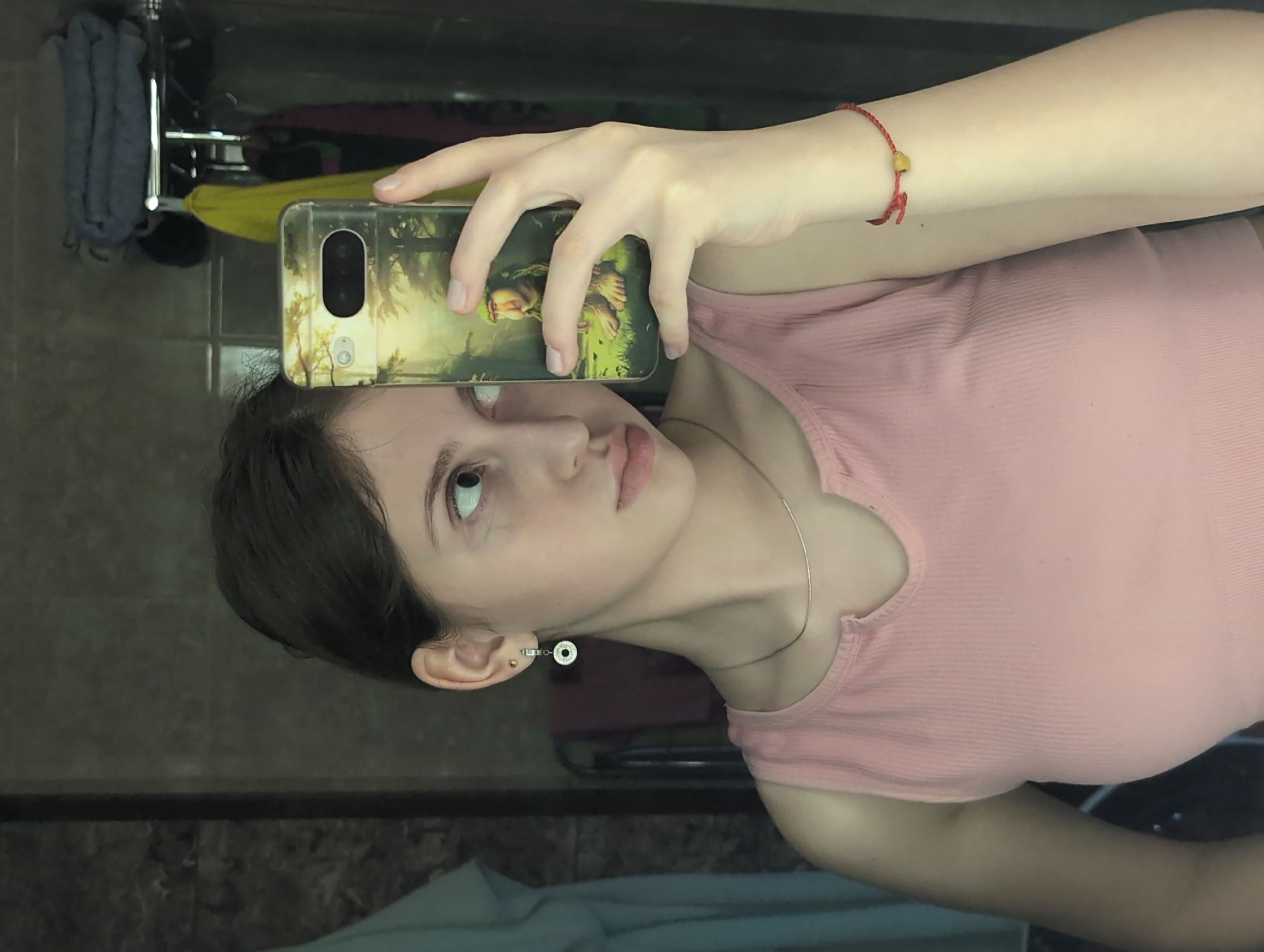 A young woman is taking a selfie in a bathroom. She is holding a phone with a case featuring a depiction of forest creatures and a landscape. She is wearing a pink tank top, a thin silver necklace, and a red bracelet with a yellow bead. Her hair is short and dark. A towel rack with gray towels is visible in the background. The location of the image is not identifiable as Izhevsk, Russia, but rather appears to be a domestic bathroom setting.