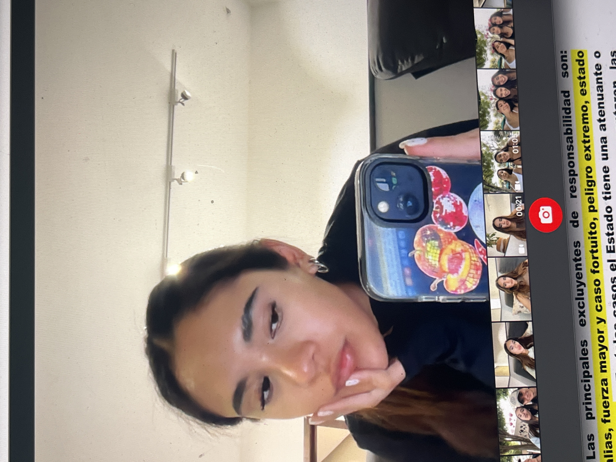 The image shows a person holding a smartphone, taking a selfie, with a screen displaying what appears to be a social media application, possibly TikTok. Multiple videos featuring the same person are previewed on the screen. Adjacent to the video previews is text in Spanish that reads, "Las principales excluyentes de responsabilidad son: alias, fuerza mayor y caso fortuito, peligro extremo, estado La casos el Estado tiene una atenuante o las." The background includes a room interior with a light fixture on the wall. The person in the image is facing the camera with a hand resting on their chin. The phone case has an apple-themed design with disco ball elements.