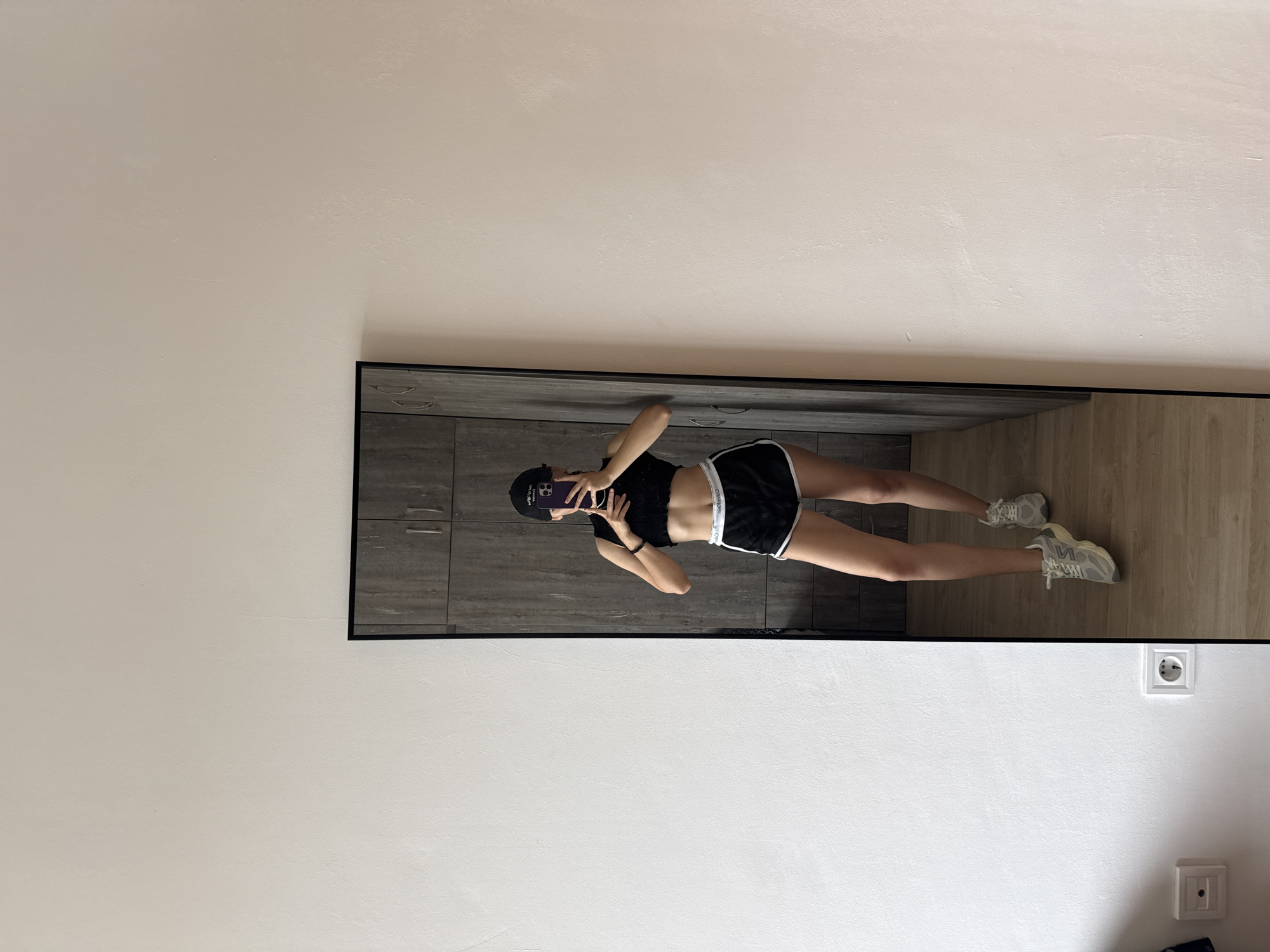 The image is a mirror selfie of a woman. She is wearing a black crop top, black shorts with white trim, a black baseball cap, and light-colored sneakers. She is holding a purple smartphone in her hands, presumably taking the photograph.
The reflection in the mirror reveals details of the room: a cabinet with a gray-toned wood-grain pattern is visible behind her, and the floor is a light wood. An electrical outlet is visible on the wall near the bottom right corner of the reflection.
The location is unknown and cannot be determined solely from the provided image, as there are no features that are unique to Plovdiv, Bulgaria or any identifiable landmarks.