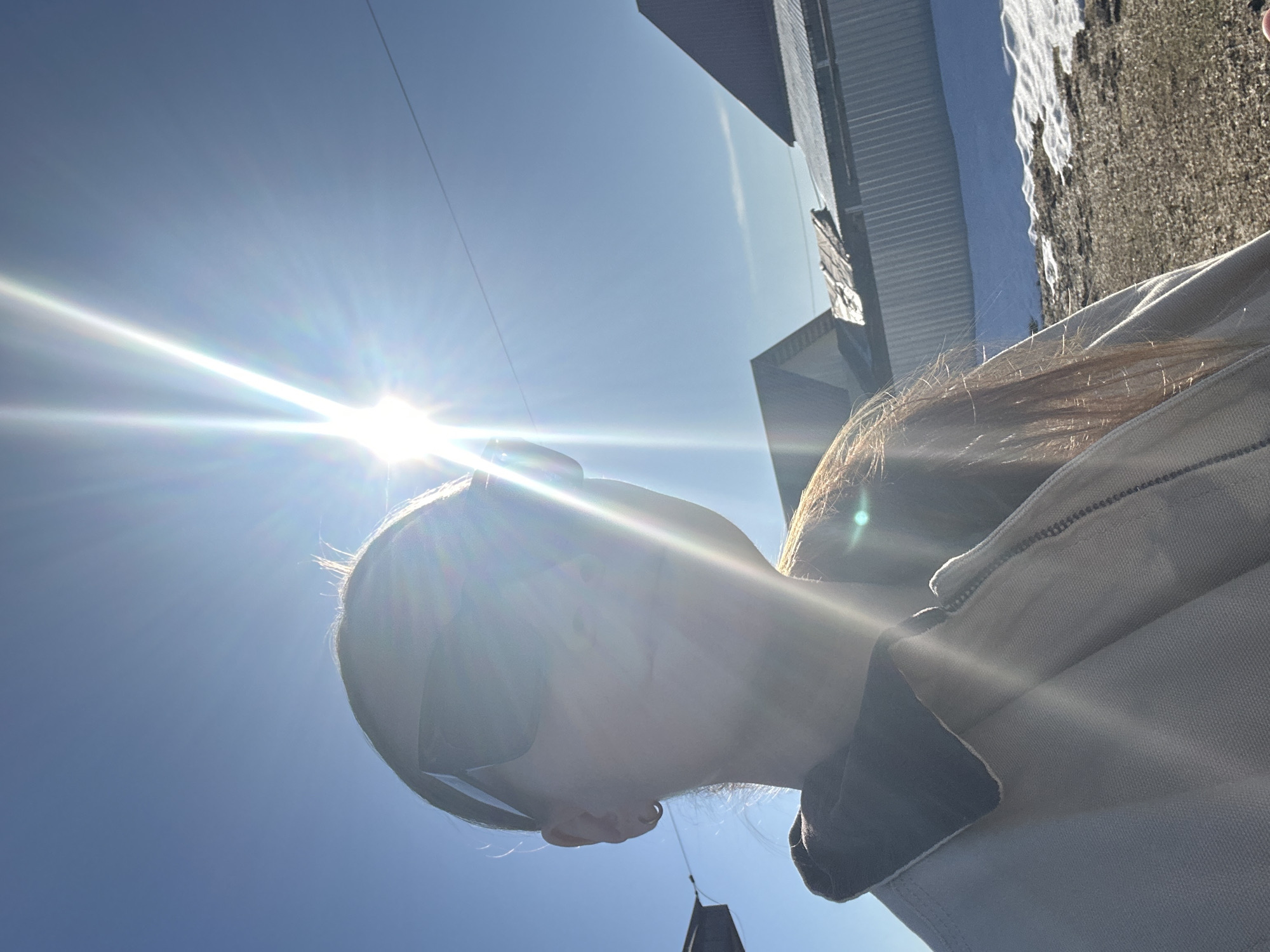 A sun-drenched, upward-facing photograph captures a person wearing sunglasses and a light-colored jacket with a dark collar. The strong sunlight obscures facial details. Behind the person, the sky is a clear blue. Buildings are visible in the background, including a structure with corrugated metal siding. A cable stretches across the top of the frame. The location is Kharnavy, Russia.