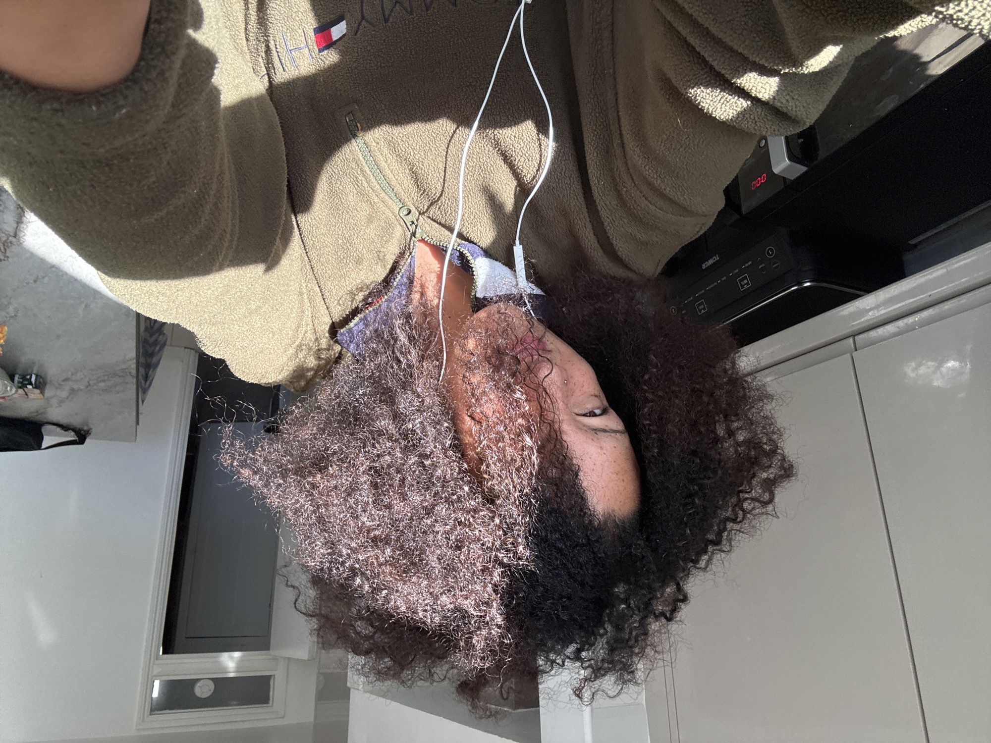 The image features a person with dark, curly hair, taking what appears to be a high-angle selfie in an indoor setting. The person's face is partially obscured by their hair, with one eye and part of their cheek, adorned with freckles, visible. They are wearing a khaki green fleece jacket with a zippered front. On the left chest area of the jacket, the text "TOMMY" is visible along with the red, white, and blue Tommy Hilfiger flag logo. White wired earphones are draped around their neck and chest.
Bright sunlight streams into the room, creating distinct shadows and highlighting the person's hair and the texture of their jacket, indicating it is daytime with clear weather. The background suggests a kitchen environment, featuring light grey cabinet doors or surfaces and a countertop with a marble-like pattern. Visible on a surface in the background are kitchen appliances: a black unit, likely an air fryer or multi-cooker, with the brand name "TOWER" and control buttons labeled "MENU," "MEAL," "TIME," and "TEMP." Next to it is another smaller electronic device with a red digital display showing "0:00." A window frame is also visible in the background, reinforcing the indoor setting. The overall scene conveys a casual, everyday moment in a home, potentially in London, United Kingdom.