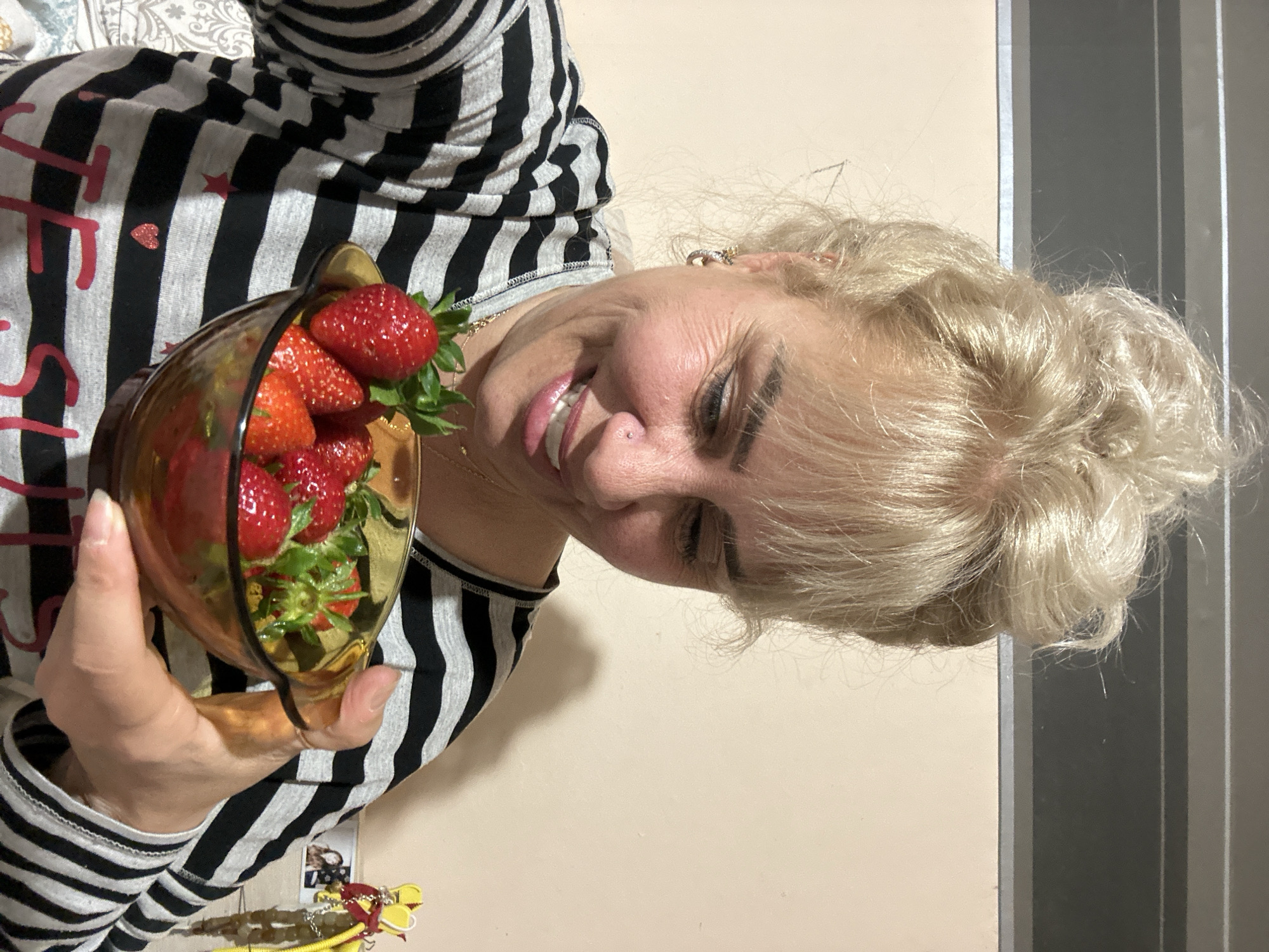 The image shows a woman holding a glass bowl filled with strawberries. She has blonde hair styled in an updo. The woman is wearing a black and white striped shirt with some red details. The background is a plain wall. There is no indication of this image being taken in Sofia, Bulgaria.