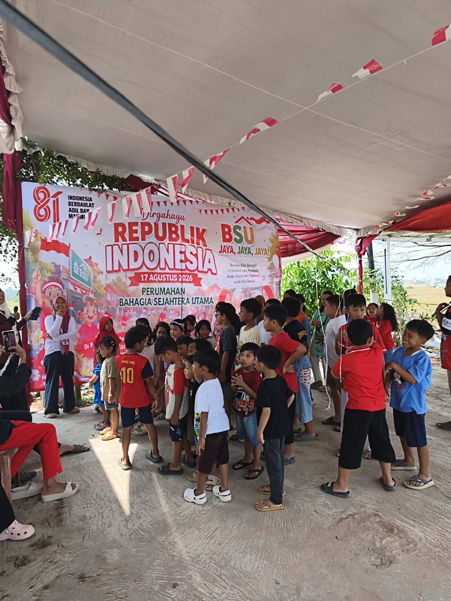 The image depicts an outdoor gathering of children and adults under a white and red tent, likely celebrating Indonesian Independence Day. A large banner in the background reads "Dirgahayu REPUBLIK INDONESIA 17 AGUSTUS 2026" and "PERUMAHAN BAHAGIA SEJAHTERA UTAMA," indicating a residential area event. The children, mostly dressed in red and white, are clustered together, some looking towards the front, while others are engaged in conversation or observation. A woman in the foreground, wearing a headscarf and a red and white patterned top, is holding a phone and appears to be taking a photo or video. The ground is a concrete surface, and the background reveals lush green trees and a bright sky. This scene likely took place in a residential complex within Bandar Lampung, Indonesia, given the text on the banner.