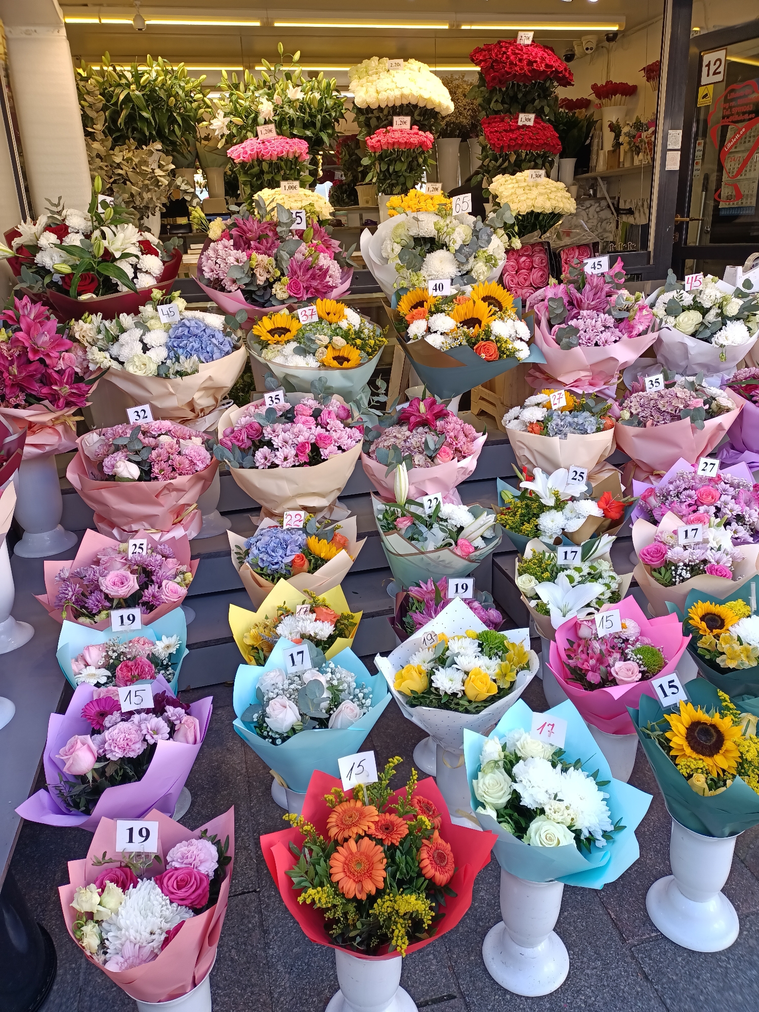 A street-level photograph shows a vibrant flower shop display in Tallinn, Estonia, with a wide variety of bouquets. The flowers are arranged in a visually appealing manner, with a mix of colors and types. Several bouquets are priced with white tags featuring numbers. The shop appears to be located on a sidewalk, with the storefront visible in the background. The lighting suggests it is daytime.