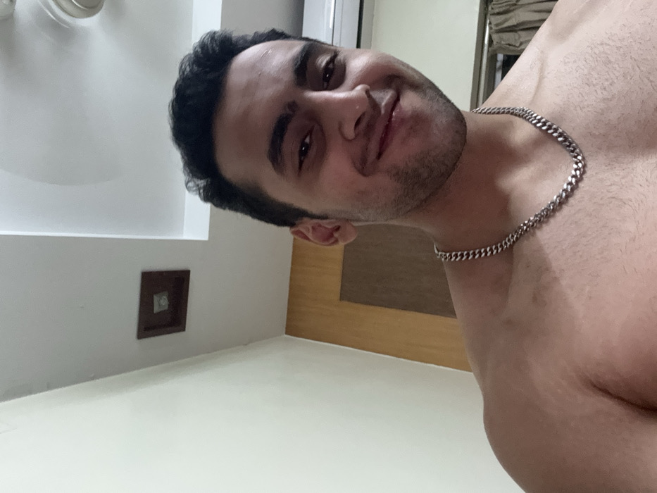 A shirtless man with a silver chain necklace smiles at the camera. He has dark hair and a slight beard. The photo is taken from a low angle. In the background is a white wall with a brown square outlet and part of a beige door. The ceiling has a light fixture. The floor is white.