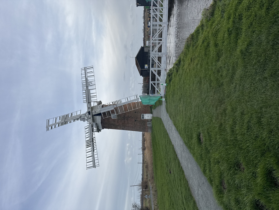The image depicts St Benet's Abbey Windmill, located near Ludham in the North Norfolk district of the United Kingdom, under an overcast sky.
The scene features a prominent brick tower windmill with a white cap and four white, lattice-framed sails, some of which exhibit missing slats, indicating disrepair or maintenance. A white rectangular door is visible at the base of the mill, and a small white-framed window is present higher on the tower. A light grey gravel path curves from the lower-left foreground directly to the mill's entrance. Green grass covers the ground on either side of the path and along the banks of a narrow canal or river running parallel to the path on the right.
A white, metal-truss pedestrian bridge spans the canal in the mid-ground. Beyond the bridge on the far bank, a dark-colored wooden boathouse or building with a dark roof is visible, along with a partially obscured second similar structure further right. A section of green mesh fencing is positioned near the windmill's base on the right side of the door. A white and orange traffic cone stands near the canal on the right, adjacent to the bridge. In the distant left background, open fields, utility poles, and a road are discernible. No individuals are present, and no actions are observed.