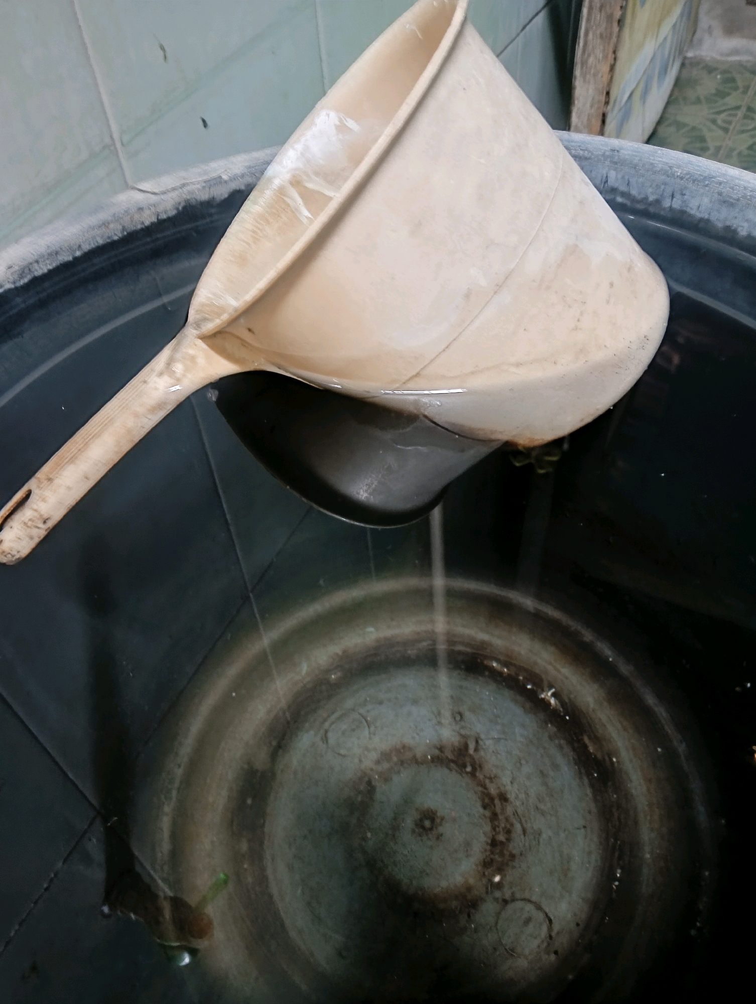 A light-colored ladle with a wooden handle rests partially submerged in dark water within a large, black tub. The ladle is positioned so its opening is angled downwards into the water, and a small amount of liquid is visible inside it. The water surface shows ripples, and the bottom of the tub is visible, appearing somewhat dirty with concentric circular patterns and scattered debris. The surrounding environment is dimly lit, with a tiled wall in the background that appears aged and somewhat grimy. The context suggests this scene is likely in a utility area or outdoor space.