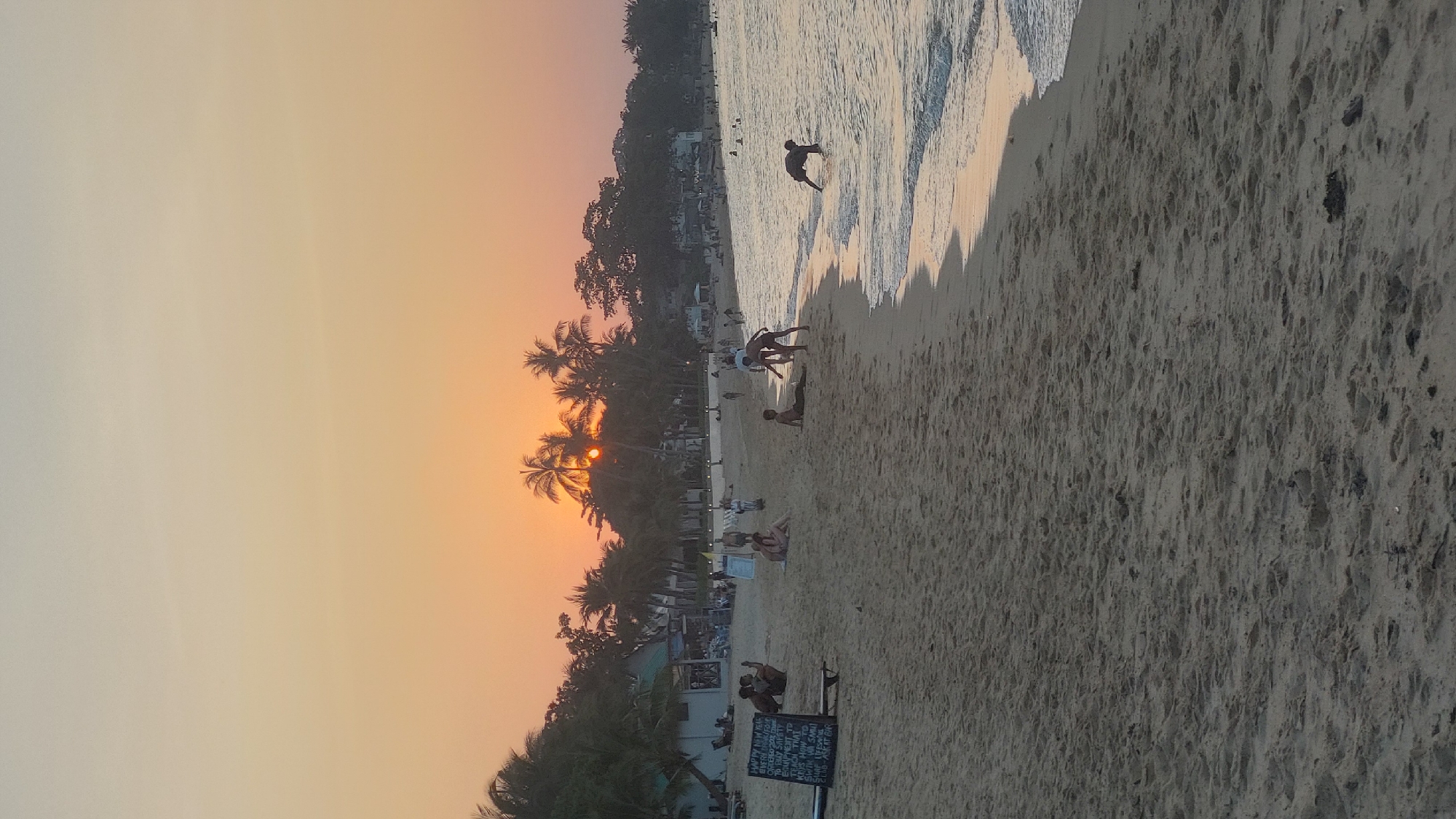 The image shows a beach at sunset in Baan Mae Nam, Thailand. The sky is a gradient of orange and yellow, with palm trees silhouetted against the setting sun. Several people are on the beach, including one person walking in the water, a group of people near the shoreline, and other individuals seated or standing on the sand. There is a menu or sign visible in the foreground. The beach is sandy, and the water is calm with gentle waves. Buildings are visible along the shoreline.