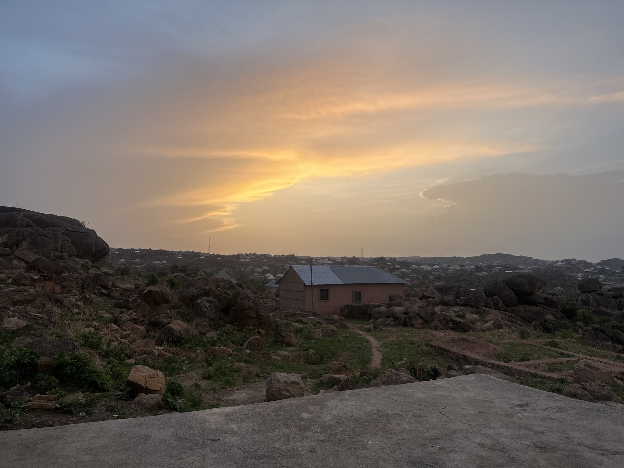 The image shows a house nestled among rocks in a landscape with a sunset in the background. The house is a simple, rectangular structure with a corrugated metal roof. There are small windows on the side visible to the viewer. A path leads from the foreground towards the house. The surrounding area is rocky and hilly, with scattered vegetation. In the background, there are other houses and structures, suggesting a settled area. The sky is colorful with a mix of blue, gray, and yellow, indicating that it is either sunset or sunrise. Based on the landscape, rocks, and architecture, it is likely that this is in the city of Jos, Nigeria. The image contains no people.