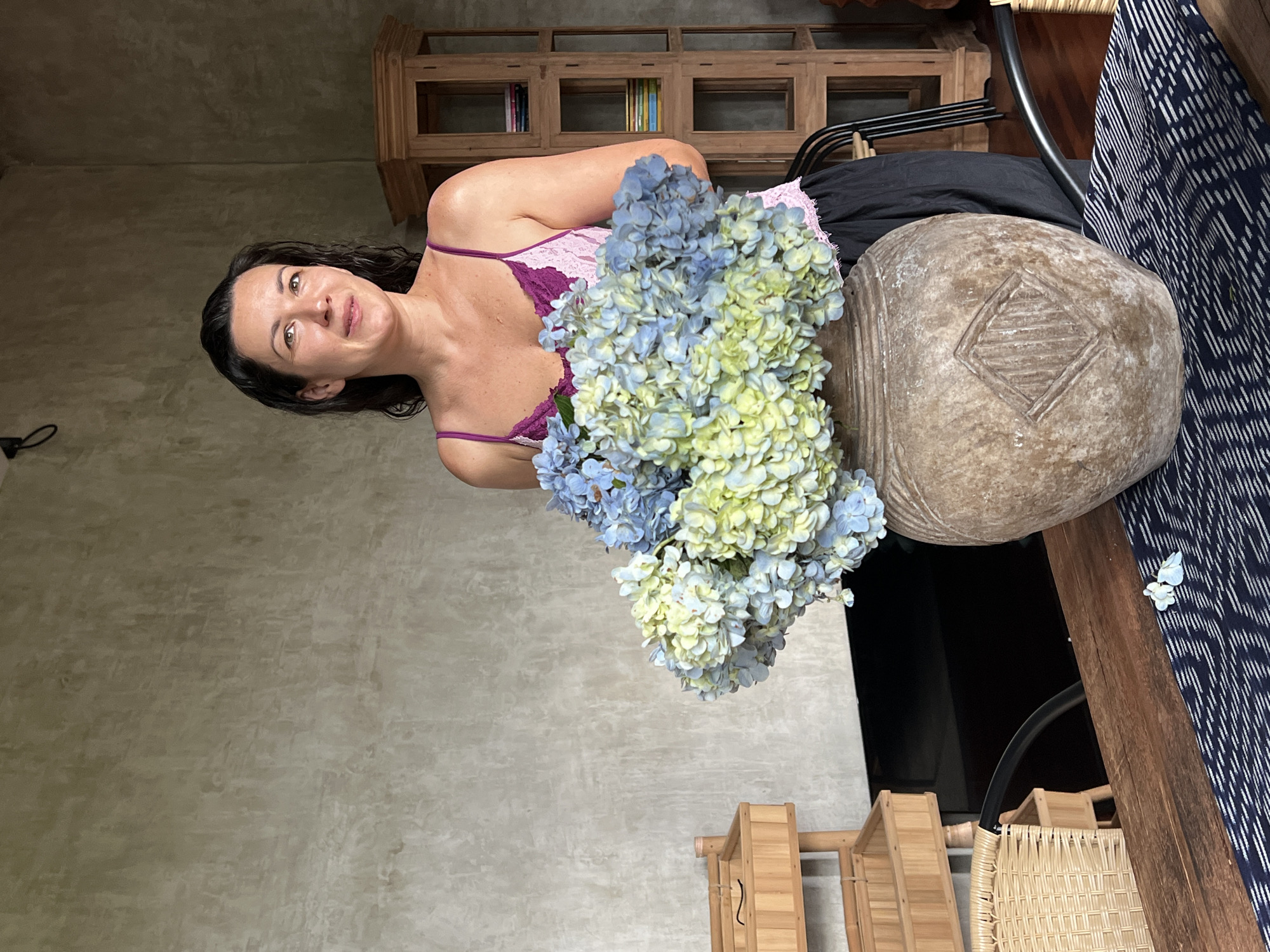 A woman wearing a purple-trimmed pink tank top poses with a bouquet of blue and yellow hydrangeas in Singakerta, Indonesia. The subject is positioned to the left of the image with a background of a light gray plaster wall. To the right is a table with a patterned blue cloth. On the table is a large, rustic, stone-like vase. Behind the subject and slightly above is a wooden cabinet. Below the table are two chairs with a woven design. A black hook is visible on the wall in the upper left corner.