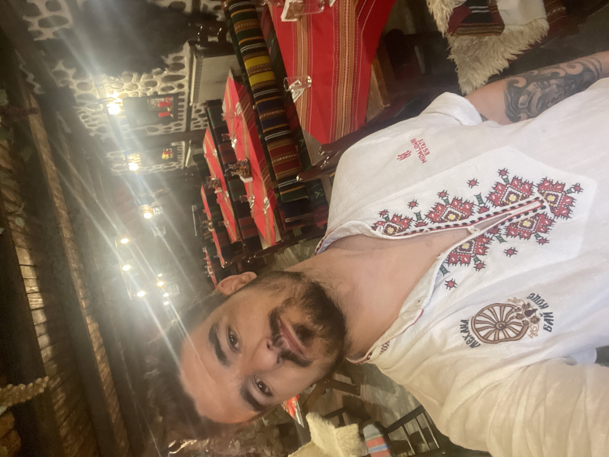 A man is captured in a selfie-style, medium shot, appearing relaxed and with a slight smile or smirk. He has dark hair, a neatly trimmed beard, and a mustache. His right forearm, partially visible, is adorned with a large, dark tattoo. He is wearing a white collared shirt featuring elaborate, colorful embroidery in red, yellow, and green patterns around the neckline and chest.
The shirt also displays two distinct embroidered texts. On the upper left chest, the word "MIDALIDARE ESTATE" is partially visible in a stylized font. On the lower right side of the chest, there is a circular emblem resembling a wagon wheel, with text curving around it. The text reads "МЕХАНА" (Mehana) on the left arc and "БАЙ МИХО" (Bai Miho) on the right arc. "Mehana" typically refers to a traditional tavern or restaurant in the Balkans.
The setting appears to be indoors, likely a traditional restaurant or tavern, suggested by the "Mehana" text and rustic decor. In the background, multiple tables are visible, each draped with red table runners over patterned, possibly woven, tablecloths featuring stripes of red, yellow, green, and black. Glasses are placed on some tables. The walls and ceiling exhibit a textured, stone-like appearance, illuminated by several bright light sources that create lens flares. Other decorative elements include furry, light-colored textiles, possibly sheepskin, and dark wooden beams. The overall ambiance suggests an evening or nighttime setting.
