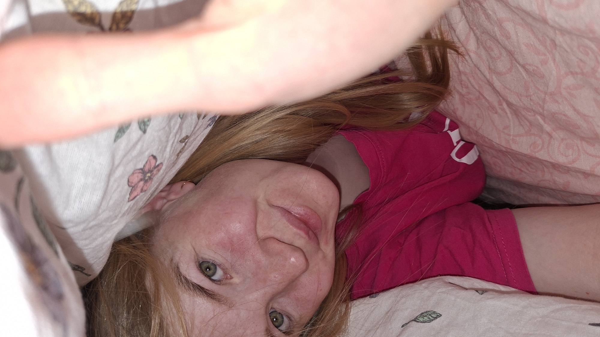 A close-up, intimate shot features a person, likely female, lying down and looking directly into the camera, suggesting a selfie or relaxed portrait. She has reddish-blonde hair, green eyes, and fair skin with visible freckles and a slight flush. A gentle, relaxed smile is present on her face, conveying contentment. She wears a bright pink t-shirt. A small, gold-colored hoop earring is visible in her right ear.
She is partially covered by two distinct types of bedding. On her left, the bedding is light-colored with a pattern of pink flowers, green leaves, and what appear to be brown butterflies or moths. On her right, the bedding is light pink with a subtle, swirled pattern. The composition gives the impression of being taken from a low perspective, from within the blankets, enhancing the cozy and personal atmosphere of an indoor setting, likely a bedroom. The lighting is soft and bright, with some areas appearing slightly overexposed, particularly towards the top left. No text is visible in the image.
