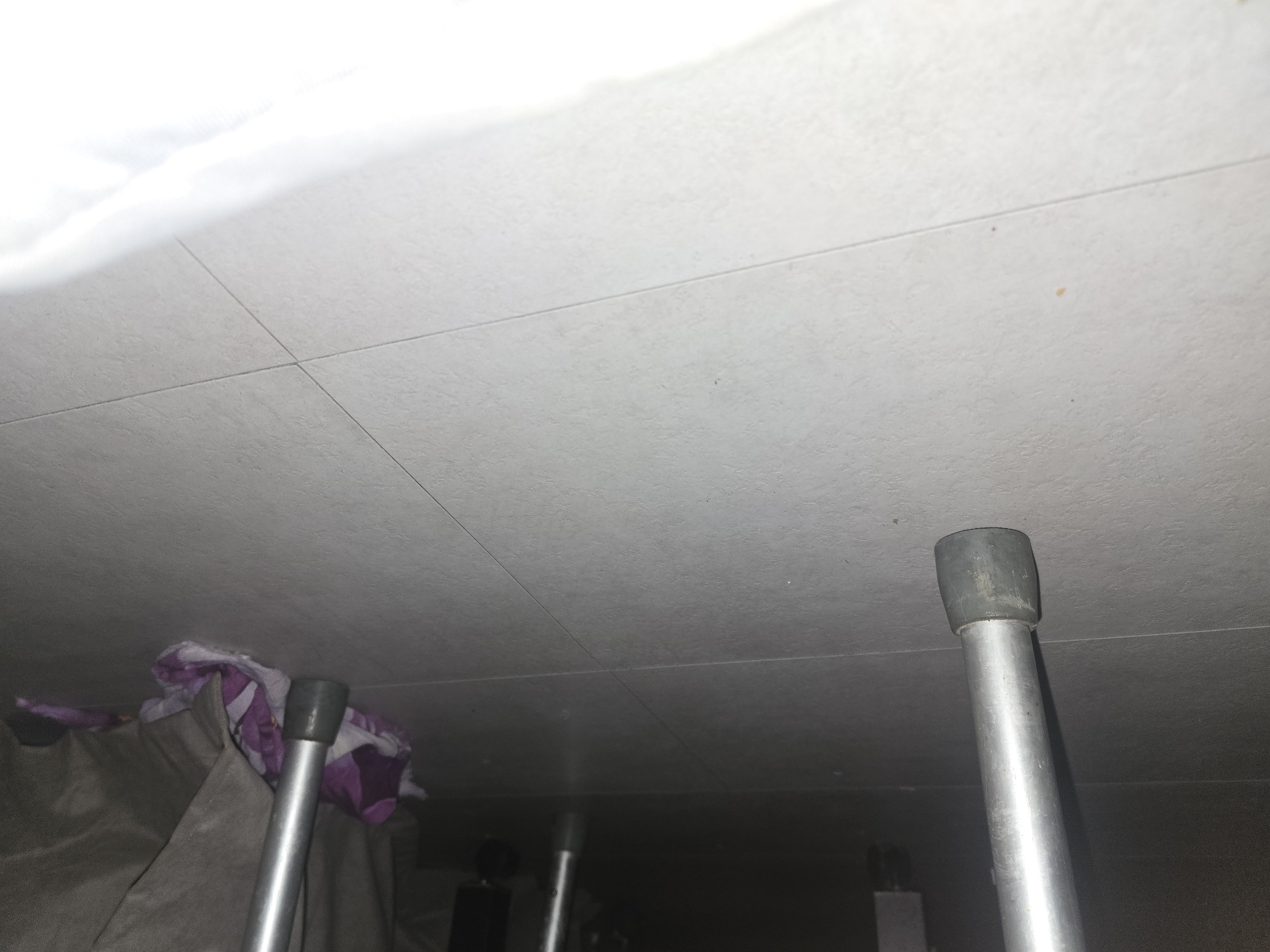 The image shows the bottom section of two silver-colored metal crutches resting on a tiled floor. The crutches have black rubber tips. To the right, a crumpled purple and white patterned fabric is visible, partially obscured by a dark gray fabric, likely a blanket or curtain. The floor is tiled with light gray tiles, showing visible grout lines. The scene is dimly lit, with the upper portion of the image being very dark. There is a bright white area in the lower right corner, possibly a reflection or another object out of focus. There is no information in the image to suggest a location in Ulan-Ude, Russia.