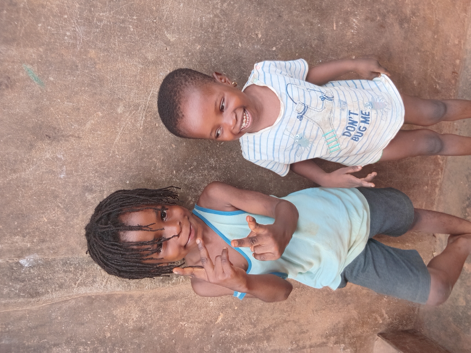 This outdoor portrait, taken in Shagamu, Nigeria, features two young children standing side-by-side on a rough, earthy-brown concrete surface. The scene is brightly lit by natural daylight, suggesting clear weather, and both children appear happy and playful as they pose for the camera.
The child on the right, who appears younger, has very short, dark hair and is wearing a white t-shirt with thin blue horizontal stripes. The front of the shirt displays cartoon illustrations of insects and the phrase "DON'T BUG ME." This child is smiling broadly, revealing teeth, and looking towards the other child with a joyful expression.
The child on the left, slightly older, has dark dreadlocks and is dressed in a light blue tank top over dark shorts. This child is also smiling, looking directly towards the camera, and is making hand gestures. One hand forms a "shaka" or "hang loose" sign, while the other hand is also performing a gesture, possibly a peace sign or similar, partially obscured.
The ground extends throughout the frame, serving as the sole background and indicating an open, outdoor setting. The overall impression is one of genuine happiness and camaraderie between the children in their environment.