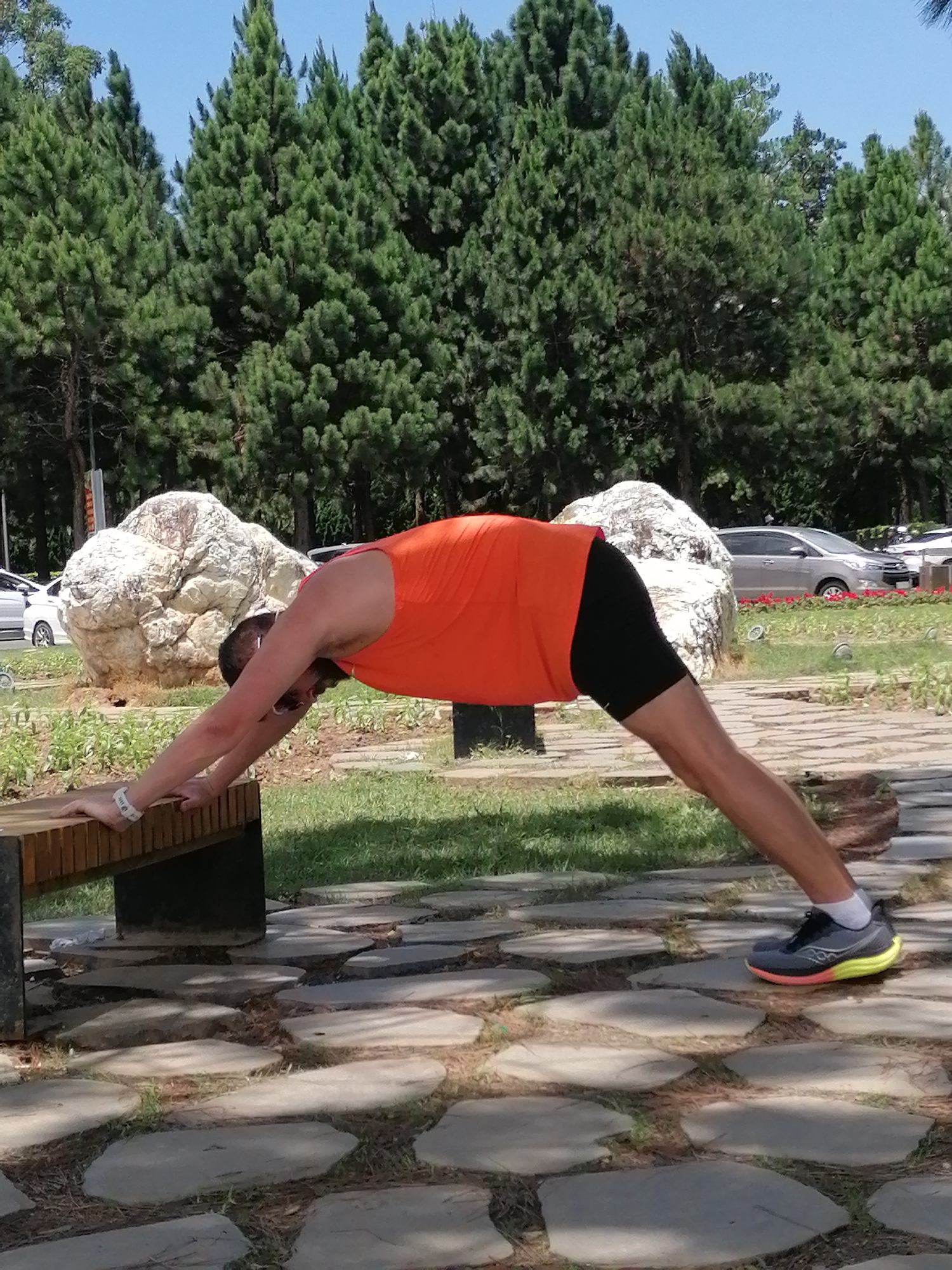 In a park in Da Lat, Vietnam, a man in an orange tank top and black shorts is performing a yoga pose, likely a downward-facing dog or a variation thereof, with his hands on a wooden bench. He is facing away from the camera, with his body extended in a plank-like position. His feet are on a stone pathway. Behind him, large white rocks and tall green pine trees are visible. A silver car is parked in the background, and a blue sky is overhead. The scene appears to be set in a public outdoor space, possibly a botanical garden or a recreational area.