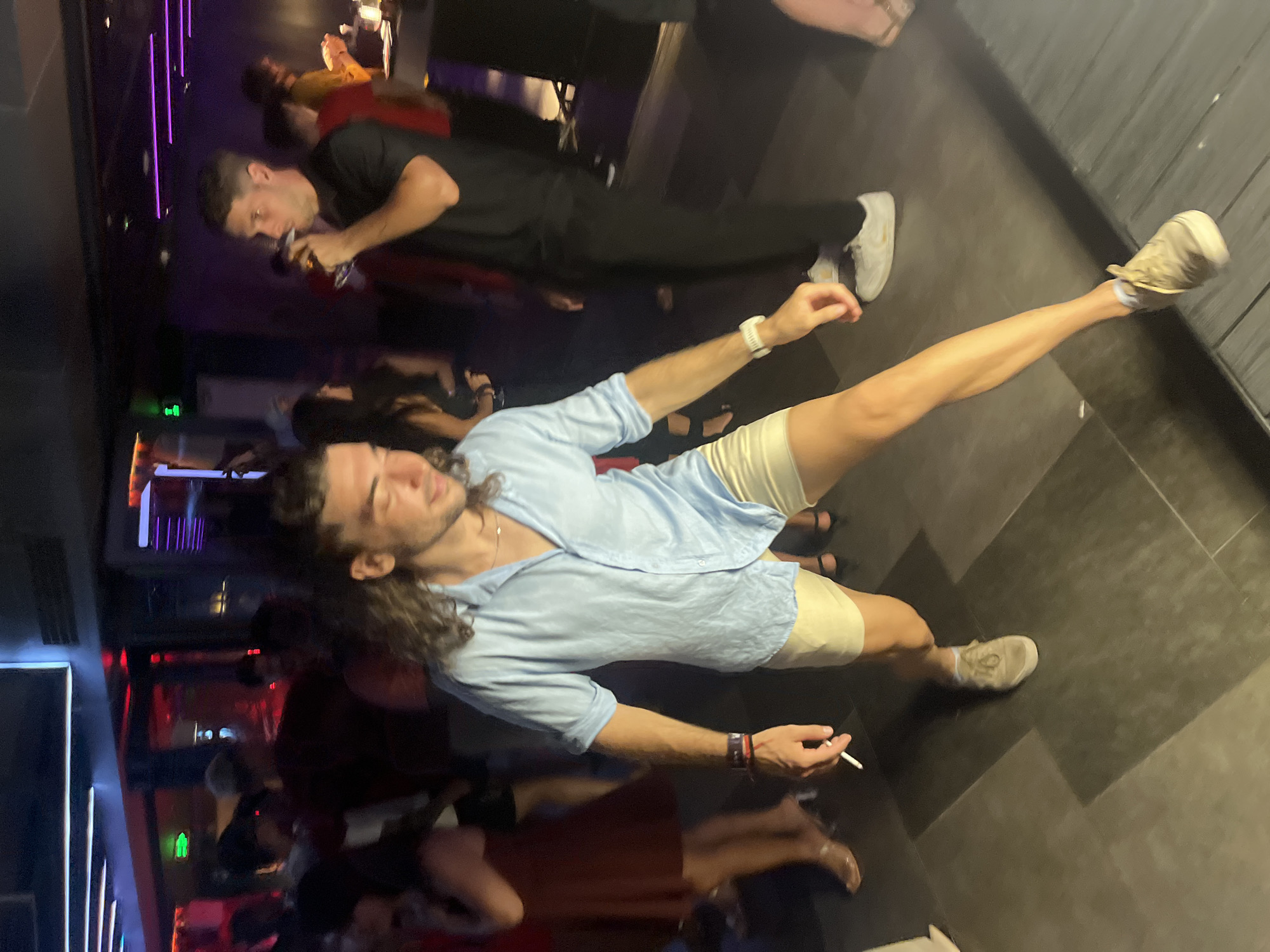 The image depicts a dimly lit nightclub or bar scene in Đà Nẵng, Vietnam. In the foreground, a young man with long, dark, wavy hair is captured mid-dance. He is wearing a light blue, short-sleeved button-up shirt and beige shorts. He is holding a cigarette in his right hand, which appears to be lit. His left hand is extended forward, and his body is in motion. He is wearing light-colored sneakers and a white watch on his left wrist. In the mid-ground, to the right, another man in a black t-shirt is drinking from a glass. Other individuals are visible in the background, some dancing and socializing, with ambient club lighting, including purple neon lights and red accents, contributing to the atmosphere. The floor appears to be tiled.