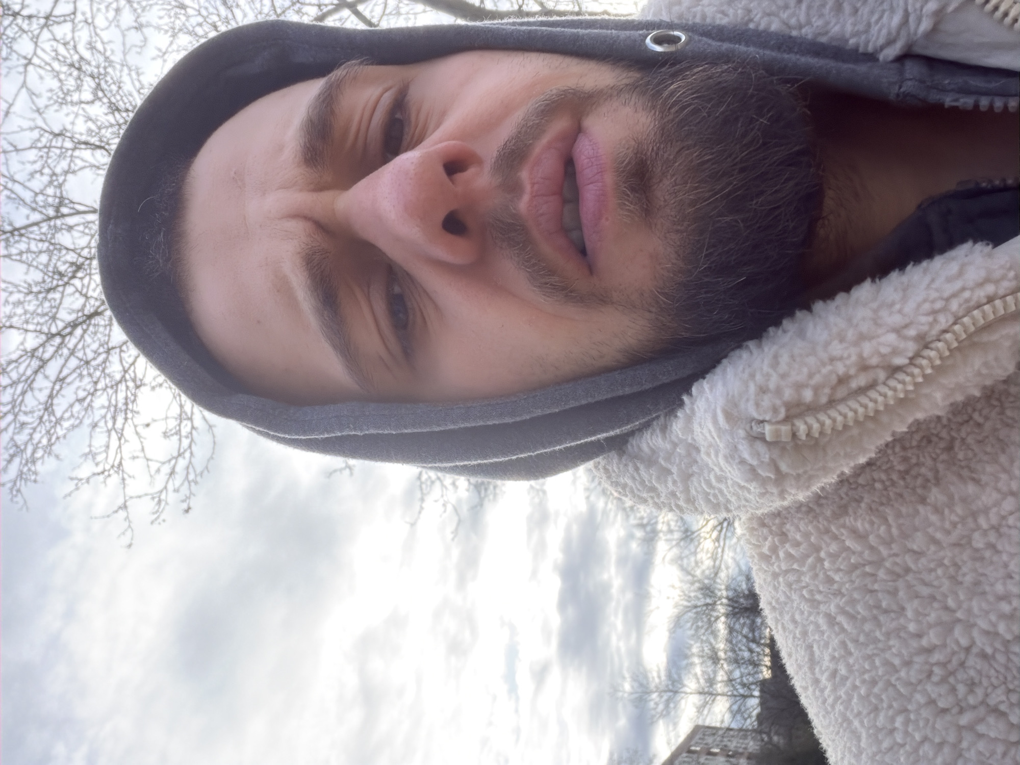 A close-up, low-angle selfie of a man wearing a grey hooded sweatshirt beneath a white sherpa-textured jacket. The man has a dark beard and appears to be grimacing or squinting. The background shows a sky with overcast clouds and bare tree branches. The image appears to have been taken outdoors. Based on the presence of bare tree branches and overcast skies, the season is likely winter or early spring. The location is unspecified and not evident from the visual details in the image. Therefore, it is impossible to place it specifically in Plovdiv, Bulgaria.