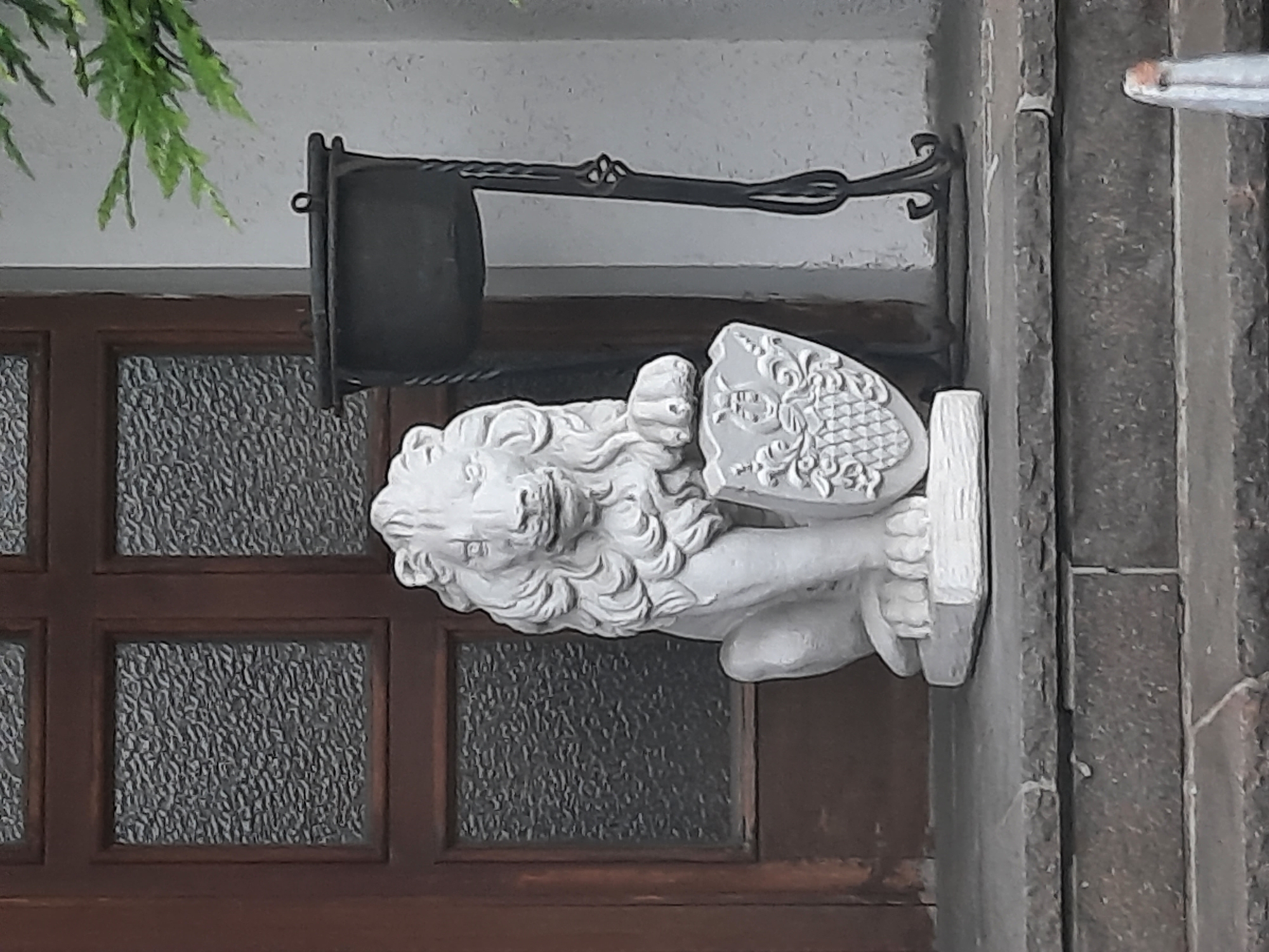 The image shows a stone statue of a lion holding a shield, positioned on a stone ledge in front of a wooden door with frosted glass panes. To the right of the lion statue is a black metal planter with a wrought iron stand. The lion statue is white and detailed, with a flowing mane and a shield bearing an ornate crest. The wooden door appears to be an entrance to a building. The overall scene suggests an exterior setting, possibly a courtyard or entryway. The image does not contain any information that would indicate it is located in Rosenheim, Germany.