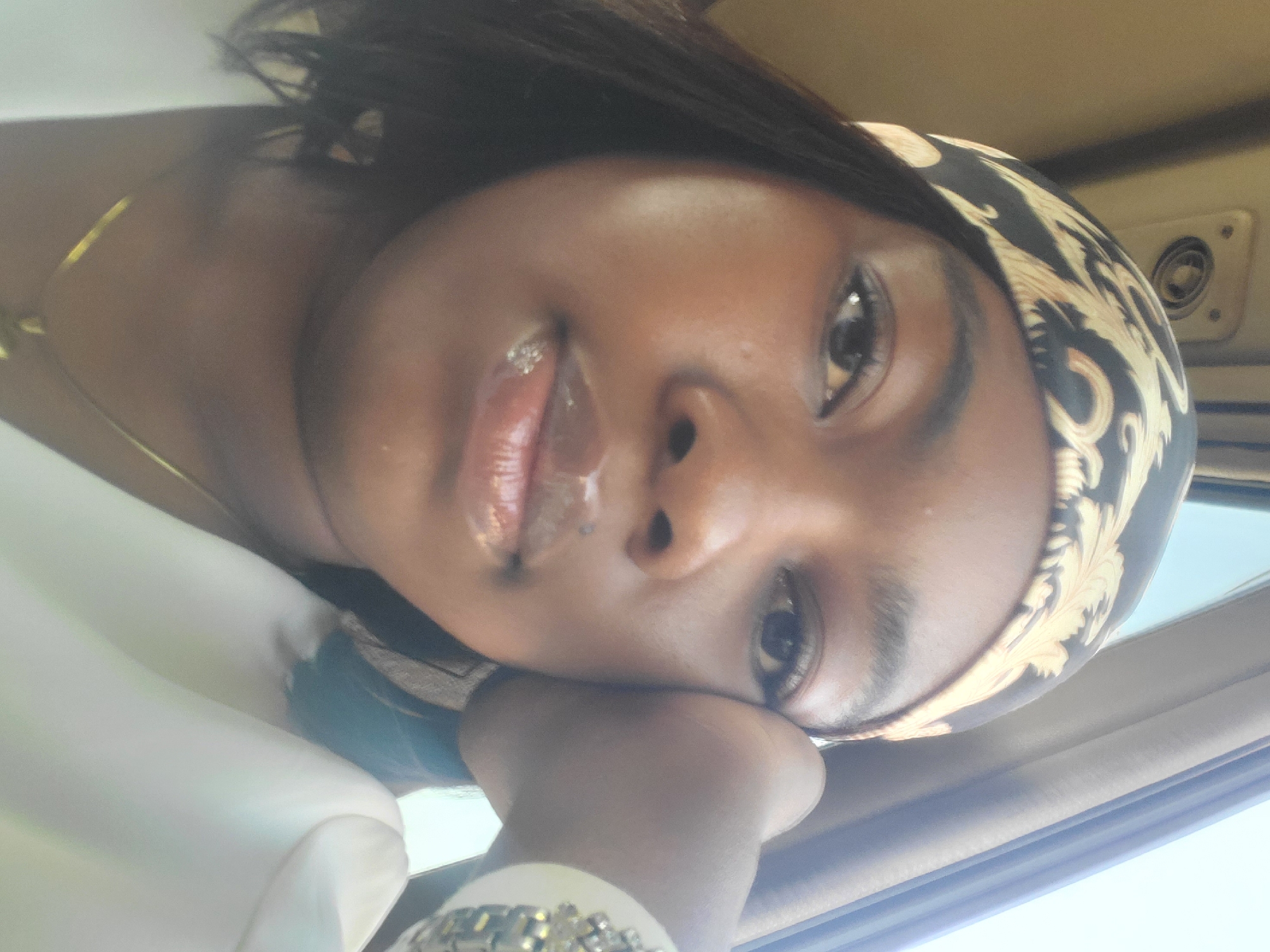 A close-up shot of a dark-skinned woman with a black and gold patterned headscarf. She is wearing lip gloss and a gold necklace. Her head is resting on her hand, which has a silver watch on the wrist. The background suggests she is inside a vehicle, possibly a car, with a white interior. There is an air conditioning vent visible on the ceiling. No specific details pinpointing Gwagwada, Nigeria, are discernible.