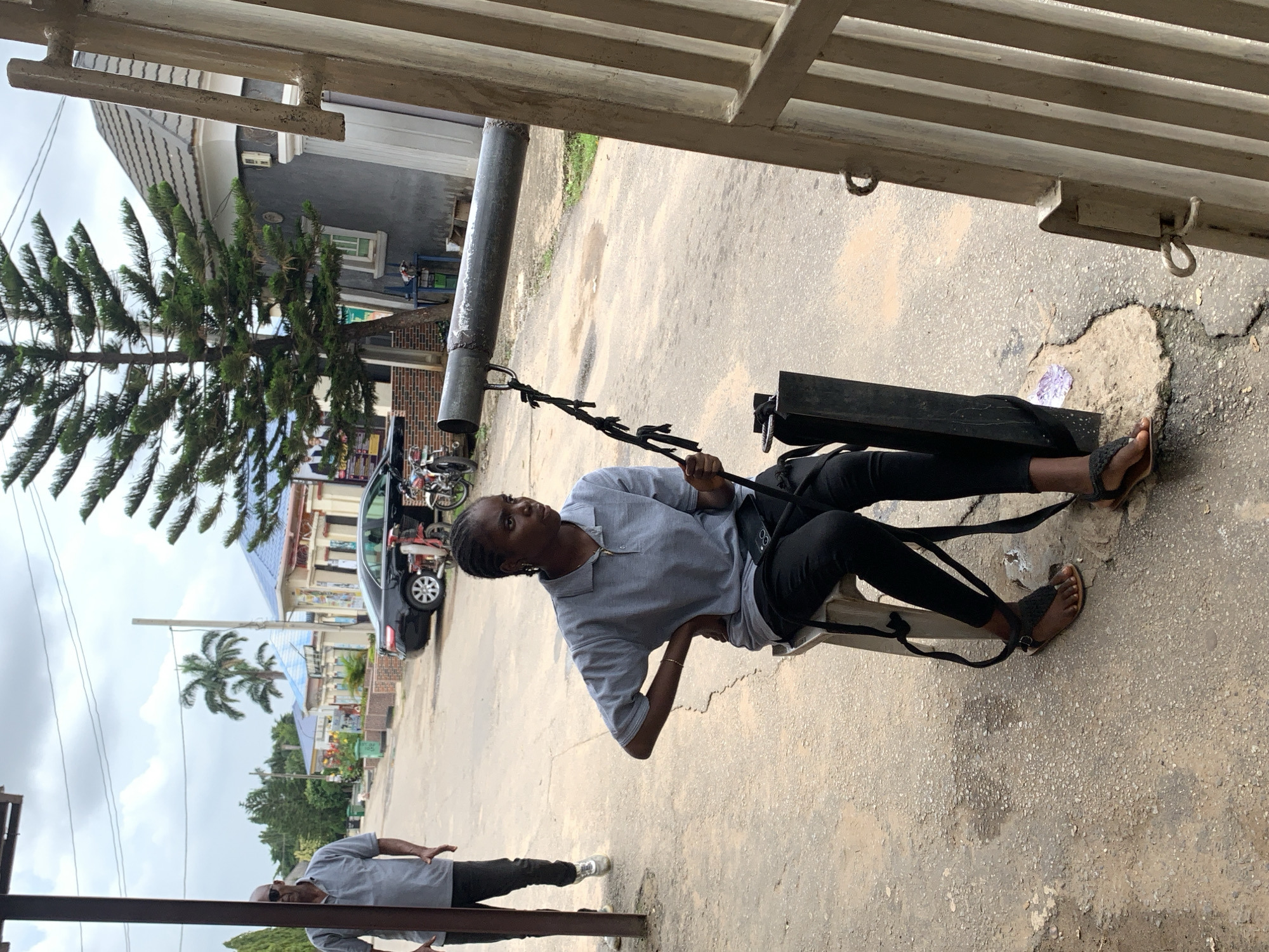 In Abuja, Nigeria, a young Black woman with braided hair sits on a plastic stool on a paved street. She is wearing a light grey polo shirt, black pants, and dark sandals. Her left hand is on her hip, and her right hand holds a dark strap that extends upwards to a metal pipe. The pipe is suspended horizontally by a rope tied to a stand. A Black man wearing a grey polo shirt and dark pants walks in the background on the left. He is bald and has sunglasses on. Several buildings with blue roofs and ornate facades are visible behind them. A black car and two motorcycles are parked on the street. The environment appears to be a residential or commercial area in Abuja.