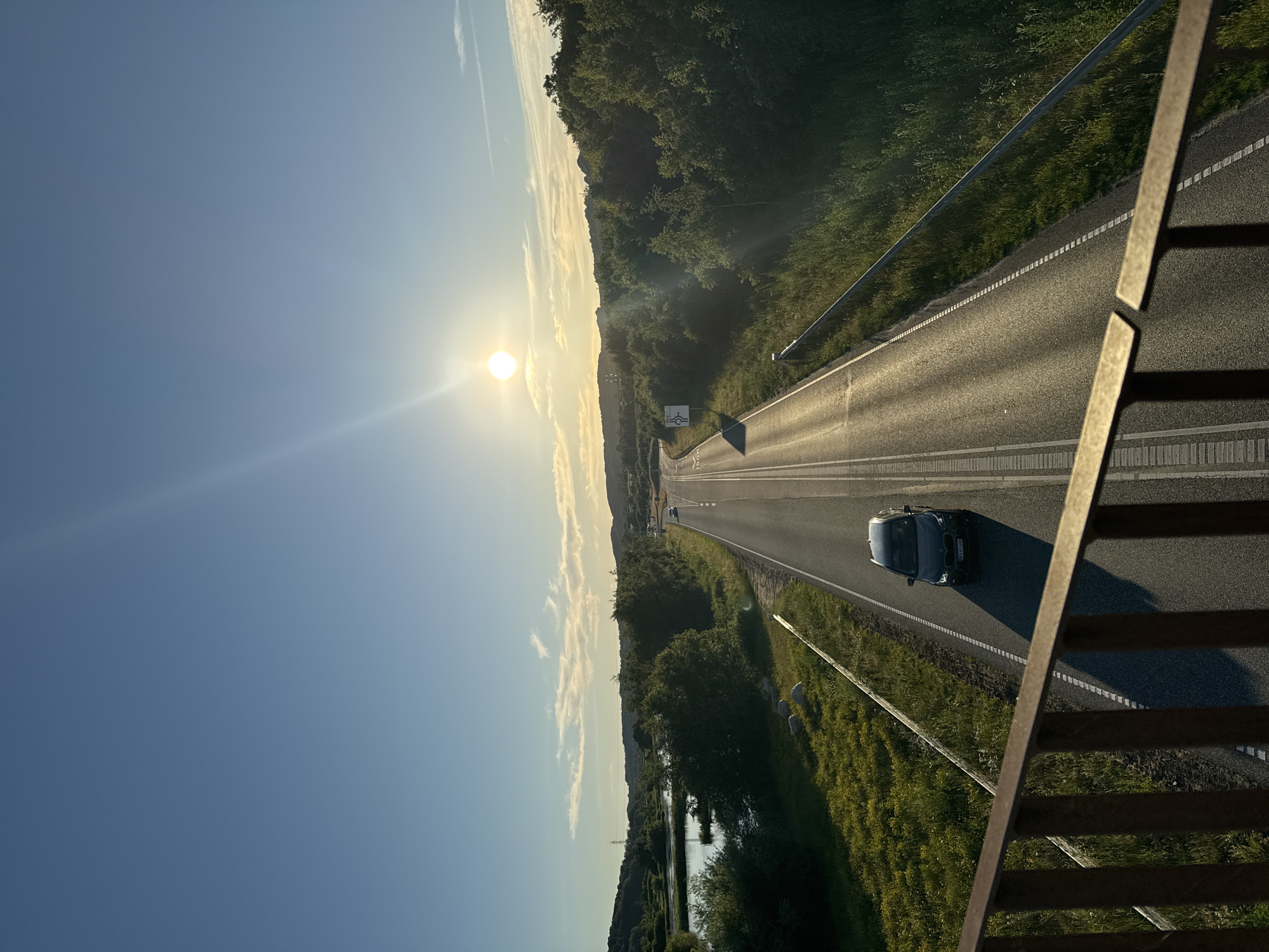 The image captures a view from an elevated position, looking down at a two-lane highway during what appears to be a sunset or sunrise. The sun is positioned high in the sky, casting a strong glare and creating lens flares. The sky is a clear blue with a few wispy clouds. The highway stretches into the distance, bordered by lush green vegetation on both sides. A dark-colored car is driving on the highway, casting a long shadow. To the left of the highway, there is a body of water, possibly a lake or river, surrounded by trees. The foreground shows a metallic railing, suggesting the vantage point is from a bridge or an elevated walkway. The overall scene depicts a scenic rural or semi-rural landscape with a road as the central element, under a bright, clear sky. The specific location in Vejle, Denmark, is not discernible from the visual elements alone, but the scene suggests a landscape that could be found in the outskirts of a city with natural elements like water and greenery.