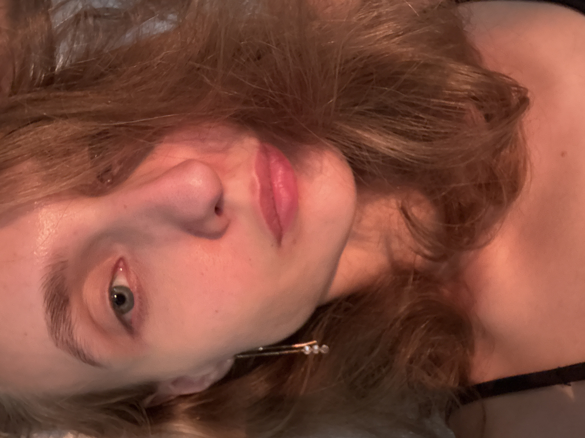 A close-up, chest-up, dimly lit photograph shows a young woman's face illuminated by warm, orange light, casting a soft glow on her skin. She has fair skin, light brown hair that partially obscures her face, and a light dusting of freckles across her nose. Her left eye, a striking blue-green, is clearly visible, while her right eye is mostly hidden by her hair. She is wearing a delicate gold earring with three small, clear stones. Her lips are parted slightly, revealing a hint of her teeth. The background is out of focus and dark, suggesting an indoor setting. The overall impression is intimate and serene, capturing a candid moment in what appears to be a personal space, possibly in or near the village of Podshivalovo, Russia, though no specific urban or rural features confirm this location.