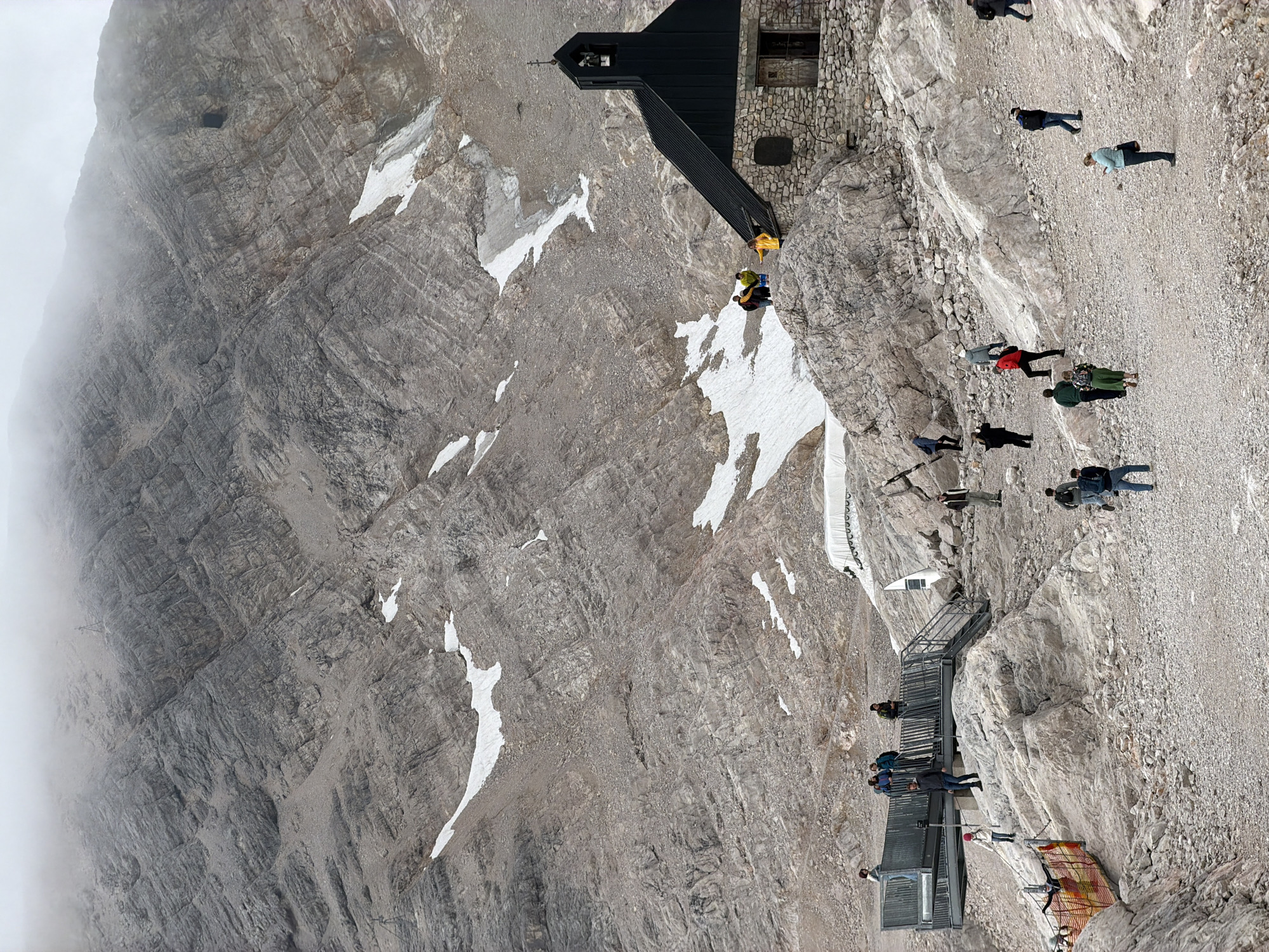 The image depicts a rocky mountain landscape with scattered patches of snow, under a hazy, overcast sky. In the foreground, a small stone building with a black roof, resembling a chapel, is situated on a rocky outcrop. A group of people are visible near the chapel, some standing on the roof and others on the rocks around it. Further to the left, a metal observation deck with stairs and railings is present, with several individuals standing on it and on the surrounding rocks. A pathway made of gravel leads through the scene, and more people are scattered on this path and among the rocks, some appearing to be walking or observing the surroundings. The mountain slopes above are steep and covered in grey rock, with more snow patches visible higher up. The overall scene suggests a tourist location in a mountainous region. This location is the Zugspitze, the highest peak in Germany, near Garmisch-Partenkirchen.