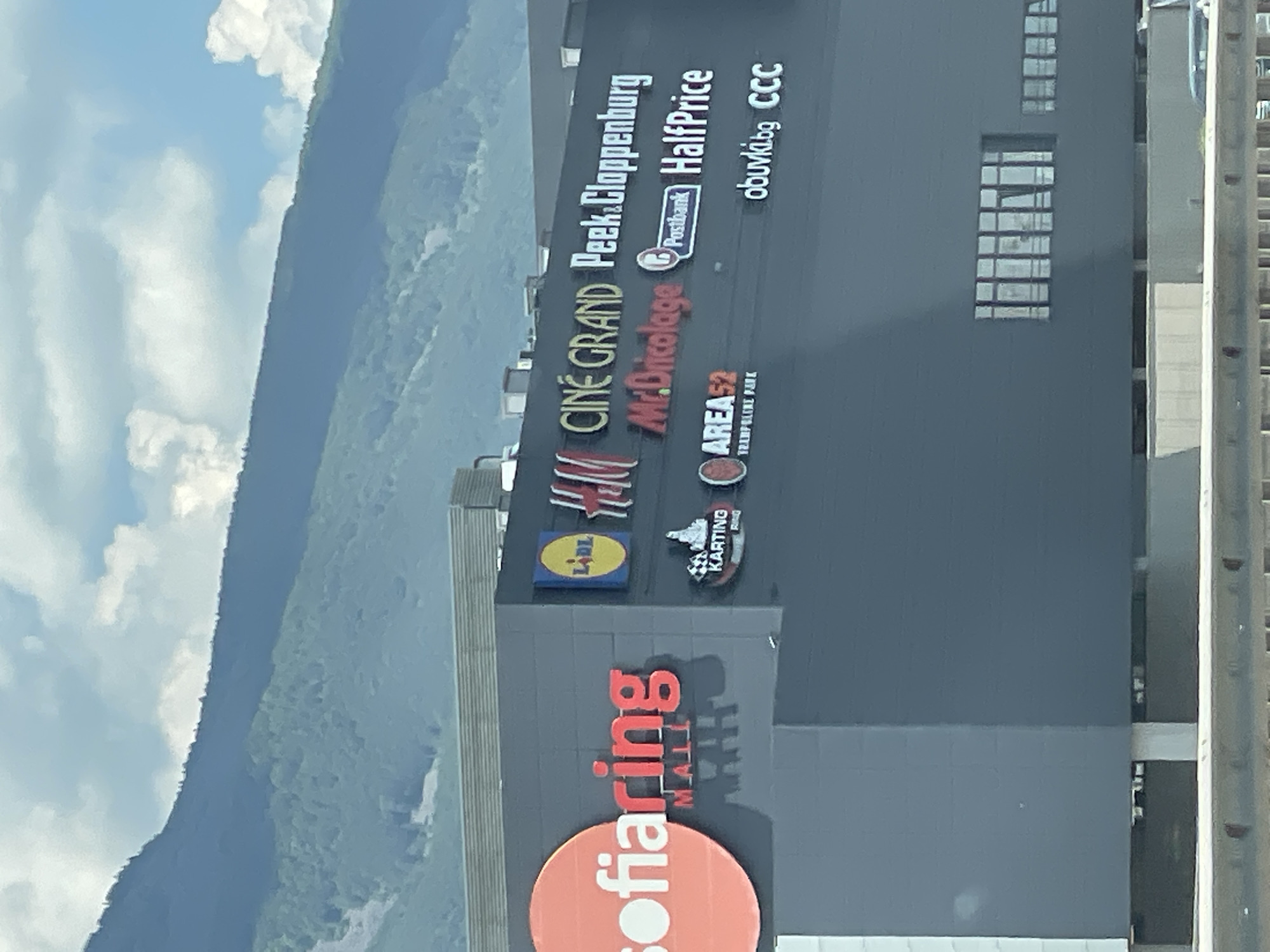The image captures a modern shopping mall with various storefronts and signage. The mall is a multi-story building with a dark grey facade. Prominent signage includes "sofiaring MALL" in large red lettering on the left, a circular orange logo with white text. To the right, there are logos and names of various businesses, including LIDL, H&M, CINE GRAND, Peek & Cloppenburg, Mr. Bricolage, Postbank, HalfPrice, AREA52 TRAMPOLINE PARK, obuvki.bg, and CCC. The mall is situated at the foot of a large, verdant mountain range, suggesting it's located on the outskirts of Sofia, Bulgaria, in an area with natural scenery. The sky above is partly cloudy with bright white clouds against a blue sky. The foreground shows a concrete barrier and part of a road, indicating the perspective is from a moving vehicle or an elevated viewpoint.