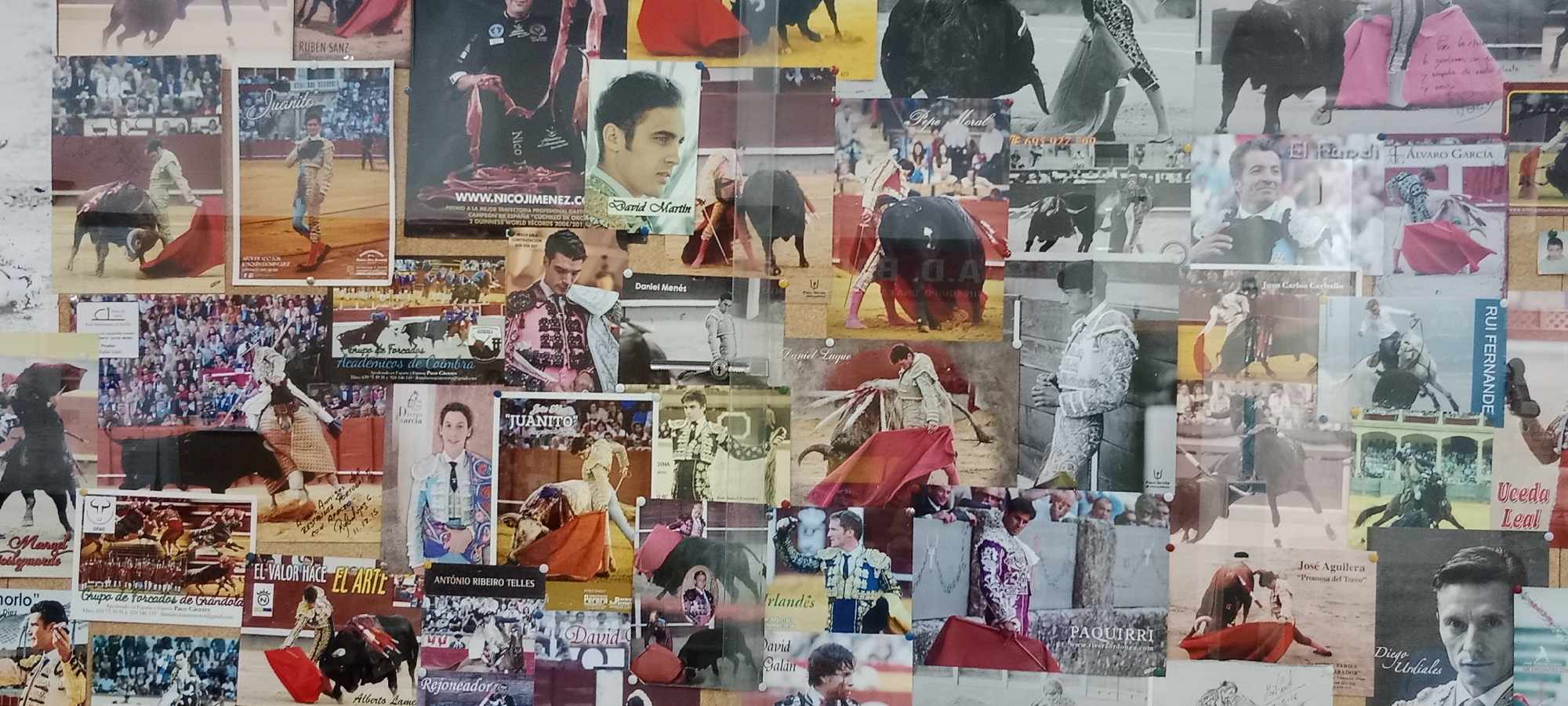 The image is a collage of posters and photographs, predominantly featuring bullfighting imagery, pinned to a corkboard. The scene is located in Almaraz, Spain, as indicated by the poster with the text "Almaraz" clearly visible in the center left of the image. The posters display images of bullfighters in traditional attire, bulls, and crowds in bullrings. Some posters include text with names of bullfighters, such as "Juanito", "Daniel Menés", "Pepe Moral", "El Randi", "Alvaro García", "Juan Carlos Carballo", "Rui Fernande", "Uceda Leal", "Jose Aguilera", "Diego Urdiales", and "Daniel Luque". There are also images of bullfighting events and group photos. The corkboard itself appears to be mounted on a wall. The overall impression is that of a display celebrating bullfighting culture, likely in a public space within Almaraz.