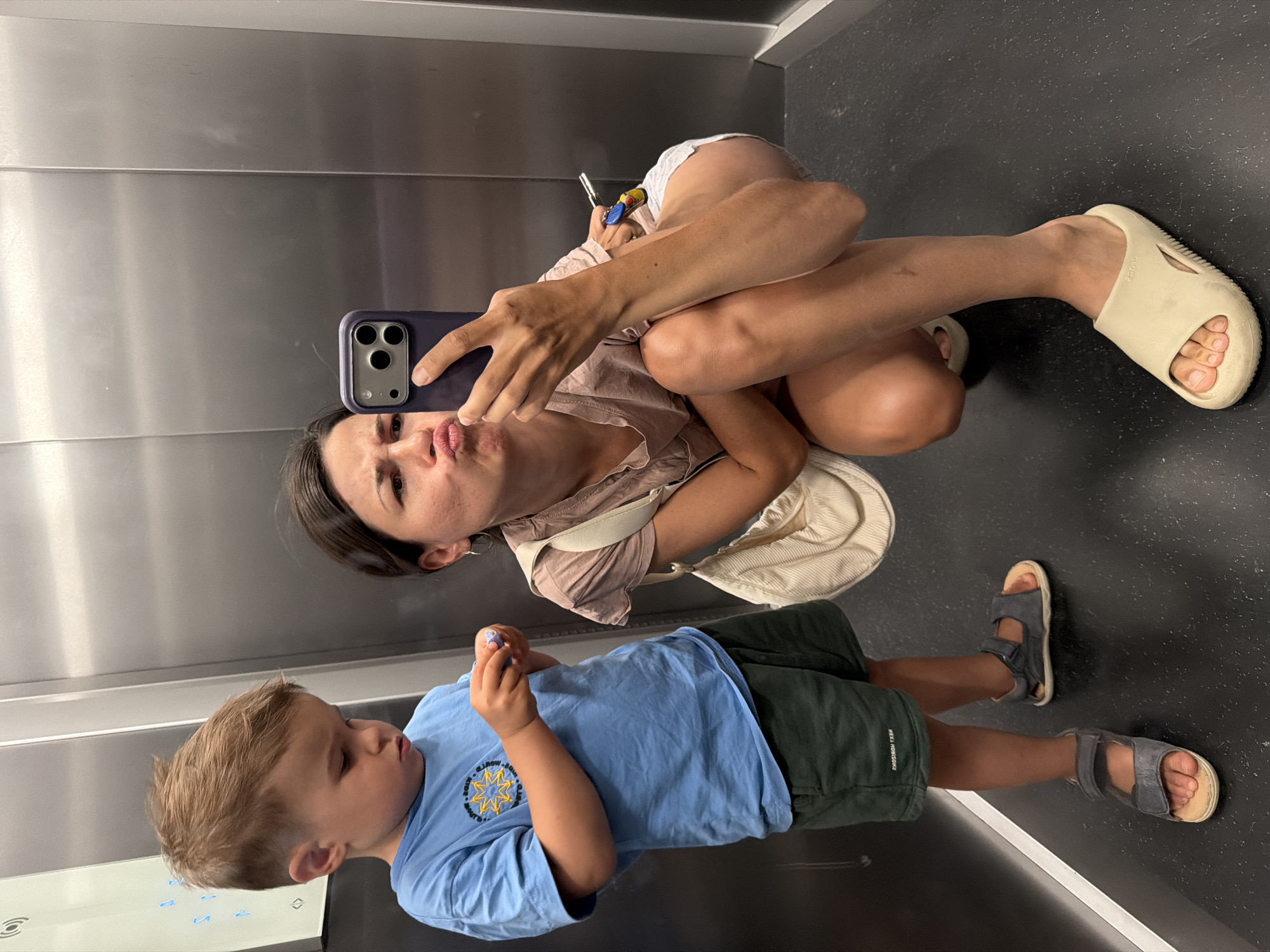 A woman and a child are in an elevator. The woman is holding a smartphone, presumably taking a selfie. She is wearing a light pink top, white shorts, and light-colored slide sandals. She has dark hair and is making a kissing face. The child is wearing a blue t-shirt with a design and dark green shorts and is looking down at something in his hands. The elevator has a metal wall. The location is unknown.