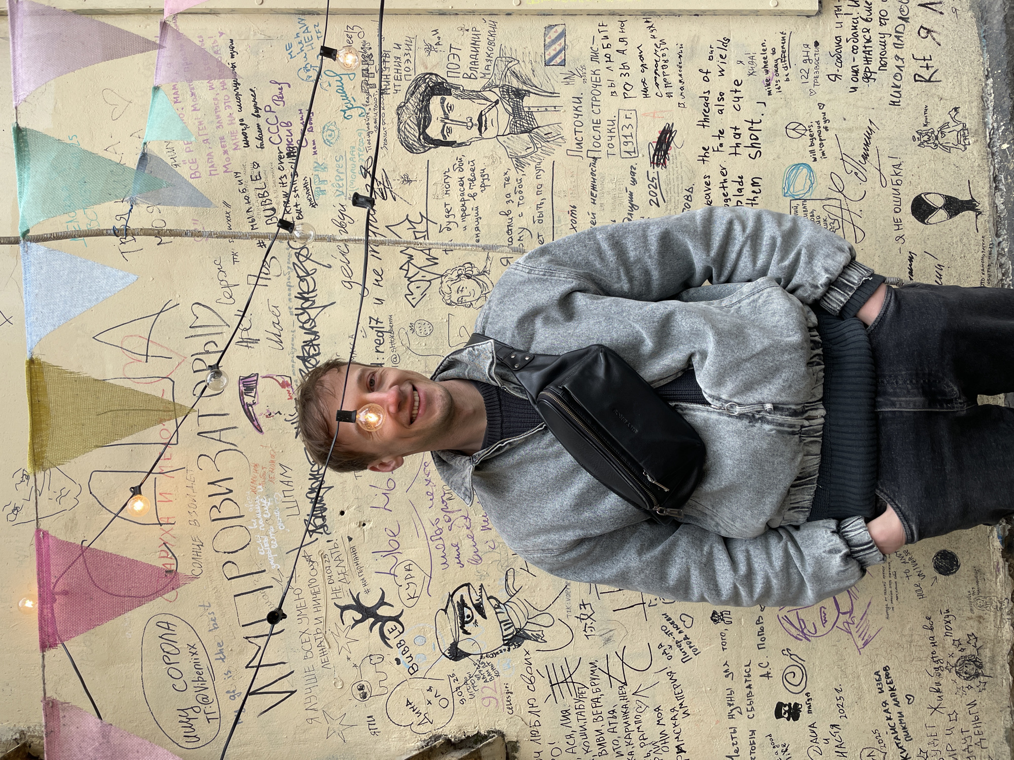 A young man poses smiling in front of a richly decorated wall covered in graffiti, text, and drawings, likely in Saint Petersburg, Russia. He is positioned slightly off-center to the left, looking directly at the viewer with a wide smile, and one of the bare string lights creates an optical illusion, appearing to emanate from his right eye.
The man is wearing a gray hooded jacket over a dark sweater and dark pants. A black fanny pack is slung across his chest. His hands are in his pockets, conveying a casual and relaxed demeanor.
The wall behind him is light yellow or beige and densely covered with various writings, drawings, and signatures in black, red, and blue ink. Above him, colorful fabric bunting in pastel pink, green, and blue triangles is strung horizontally across the wall. Numerous string lights with bare, warm-toned bulbs are strung vertically across the scene.
Notable visible text includes large Russian words like "ИМПРОВИЗАЦИЯ" (Improvisation), "Ищу СОРОЛА" (Looking for a SOUL/SOLO), and "Я ЛУЧШЕ ВСЕХ УМЕЮ ЛЕЖАТЬ И НИЧЕГО НЕ ДЕЛАТЬ!" (I can do nothing better than anyone!). There's a prominent sketch of "ПОЭТ ВЛАДИМИР МАЯКОВСКИЙ" (Poet Vladimir Mayakovsky) with associated text about "Листочки. Поэзия. Любовь. 1913г. 2025". English phrases such as "will byers. i'm so proud of you" and "it's okay to be different" are also visible. Drawings include abstract faces, a spider-like figure, and various doodles. Many personal messages, names, and dates (e.g., 2025, 04.07.25) are scrawled across the surface, reflecting a public space for expression. The overall atmosphere is lively and artistic.